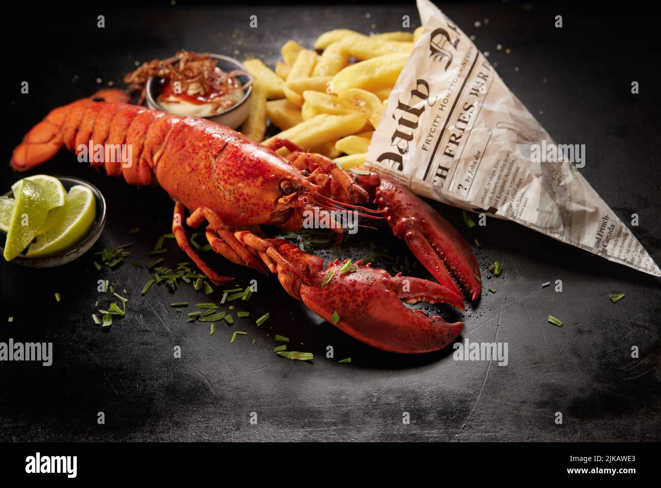 From above tasty red lobster and potato chips in newspaper sheet wrapping served on black table near lime pieces and sauce Stock Photo