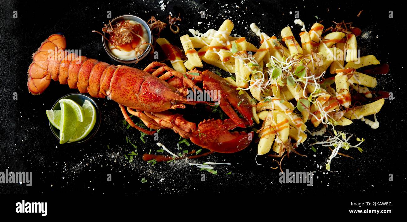 Top view of fresh yummy lobster and potato fries served on black surface near lime slices and bowl of sauce Stock Photo