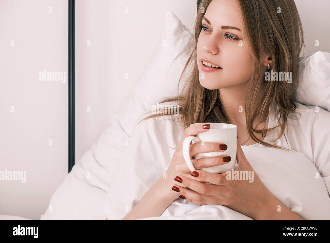 Young attractive woman lies in bed after waking up with white cup in her hand looking out the window. Morning routine, time for yourself Stock Photo
