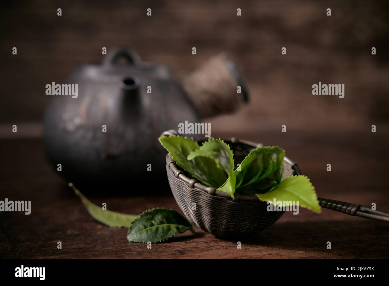 Fresh green tea leaves in ladle placed on table near old fashioned metal teapot on blurred background in light room Stock Photo