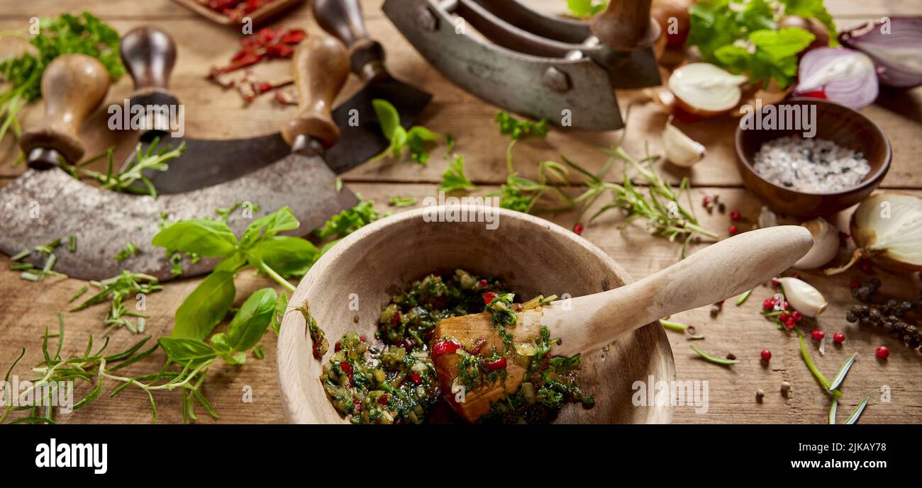 From above of wooden mortar with greens served on table with old fashioned chopping knives and scattered herbs in light kitchen Stock Photo