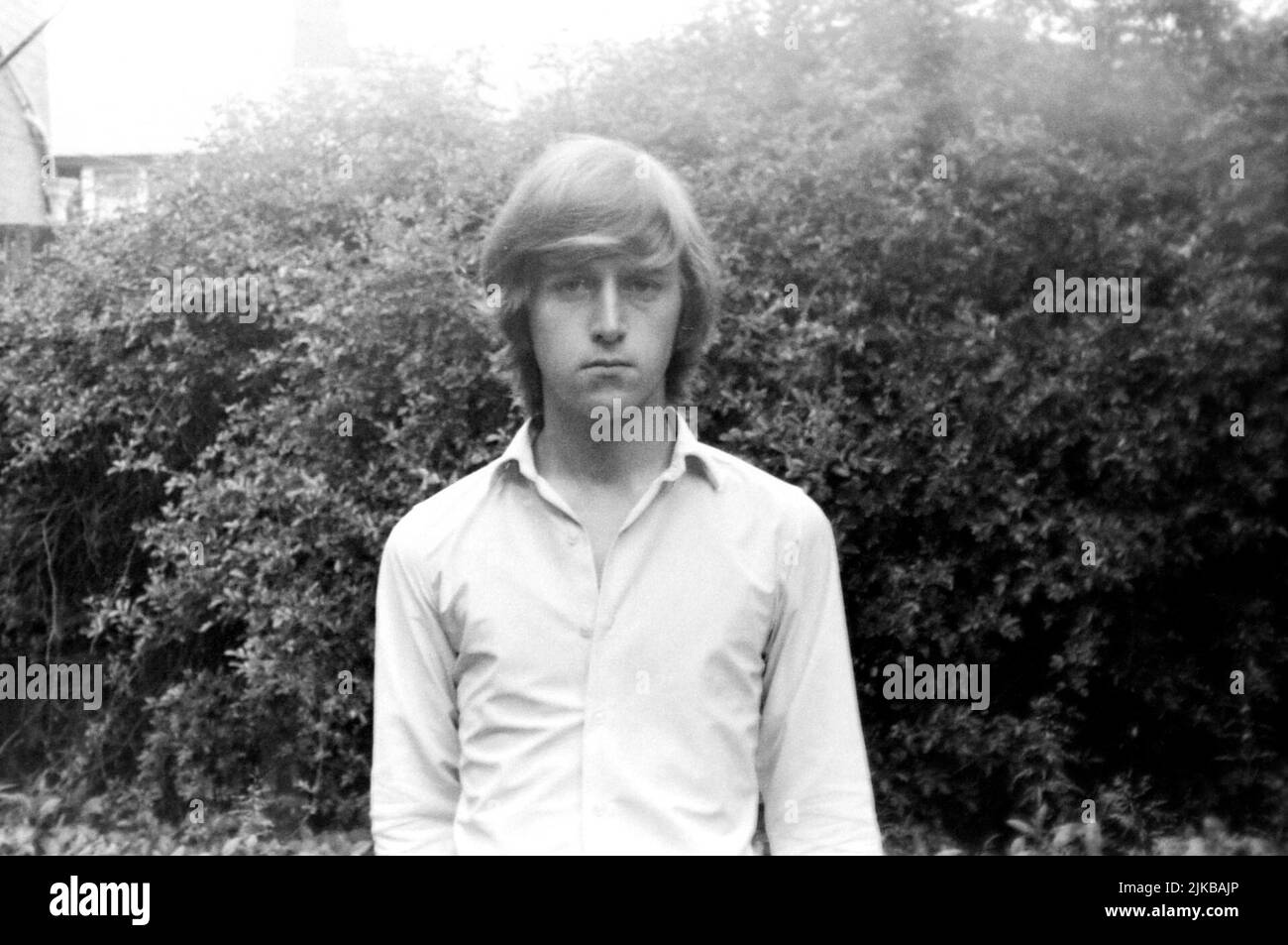 Portrait of a serious looking Caucasian teenage boy, aged 18, with long hair, taken in 1970 in a back garden with a hedge in the background. Taken in Blackpool, Lancashire, UK. Stock Photo
