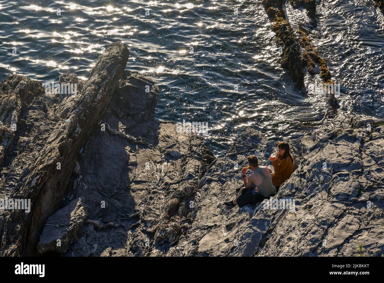 A young couple takes a snack break sitting on the rocks by the sea at sunset, Nervi, Genoa, Liguria, Italy Stock Photo