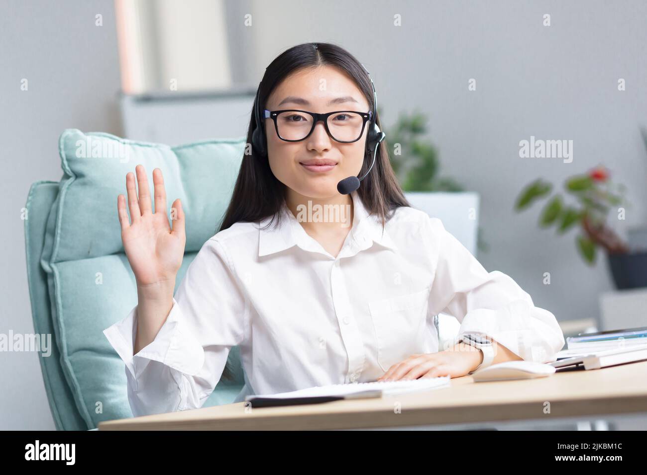 Portrait of a young beautiful Asian woman in glasses and headphones with a microphone, sitting in the office at the desk. He greets, waves his hand, conducts an online webinar Stock Photo