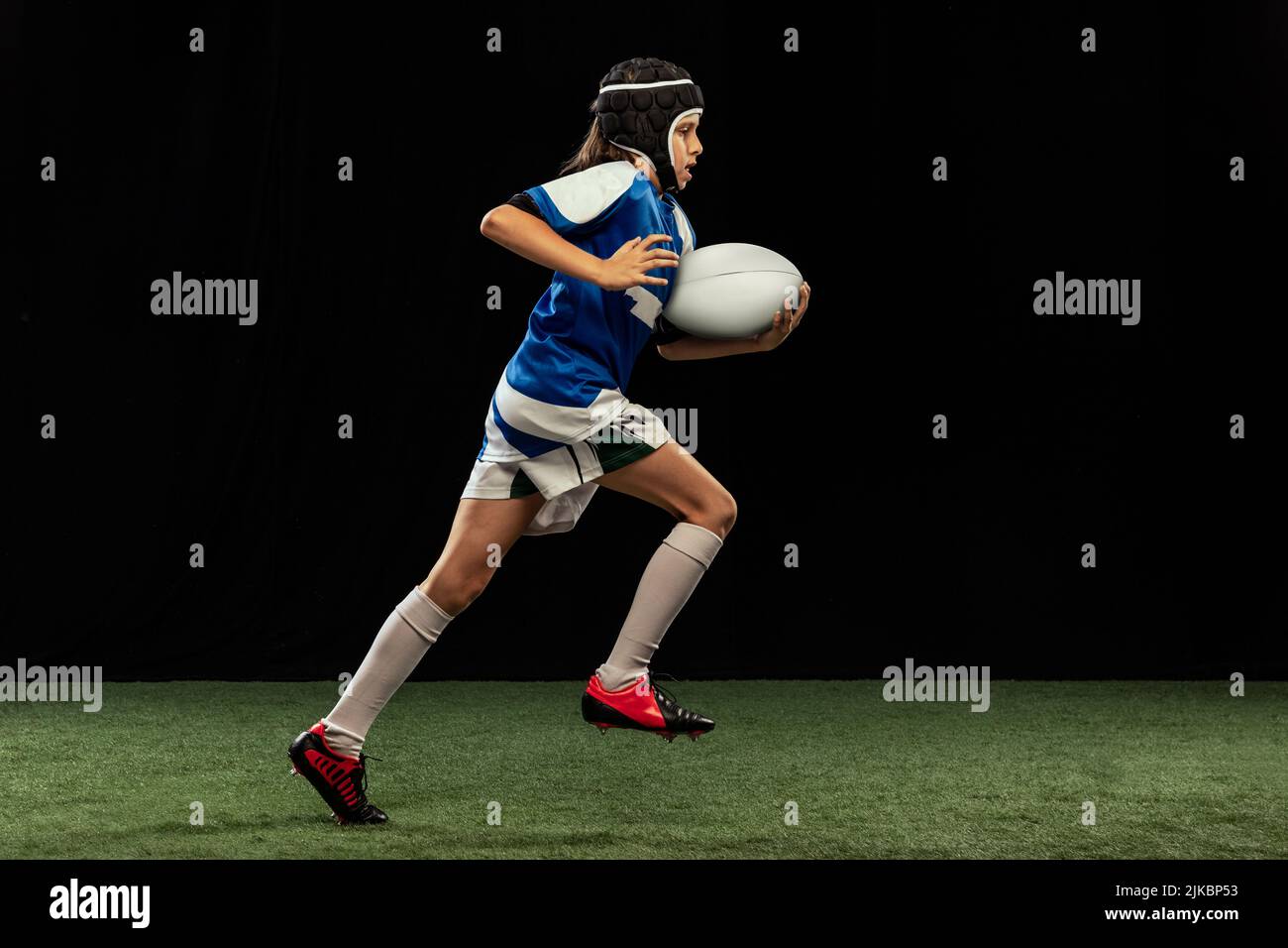 Dynamic portrait of school age boy, junior male rugby player practicing rugby football isolated on dark background with grass floooring. Stock Photo