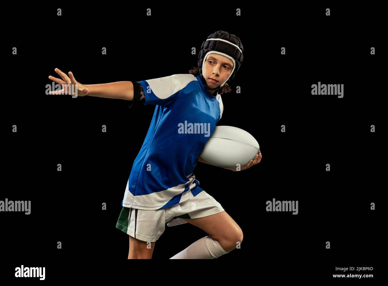 Dynamic portrait of school age boy, junior male rugby player practicing rugby football isolated on dark background with grass floooring. Stock Photo