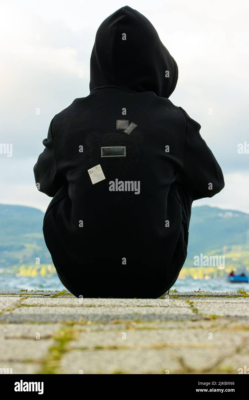 A vertical rear view of a mysterious boy in a black hoodie sitting on ...