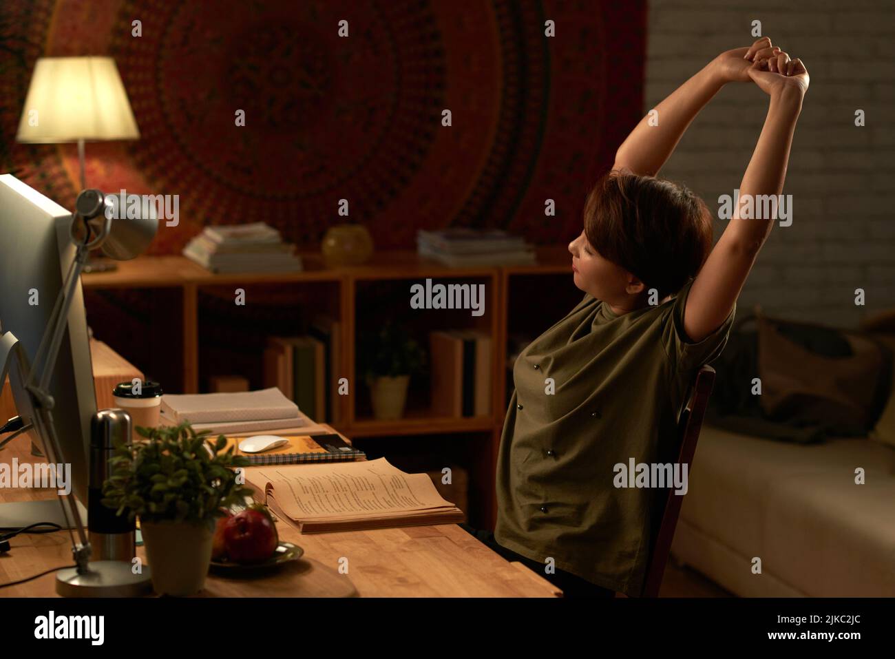 Tired college student stretching while doing homework in dark room in the light of lamp Stock Photo