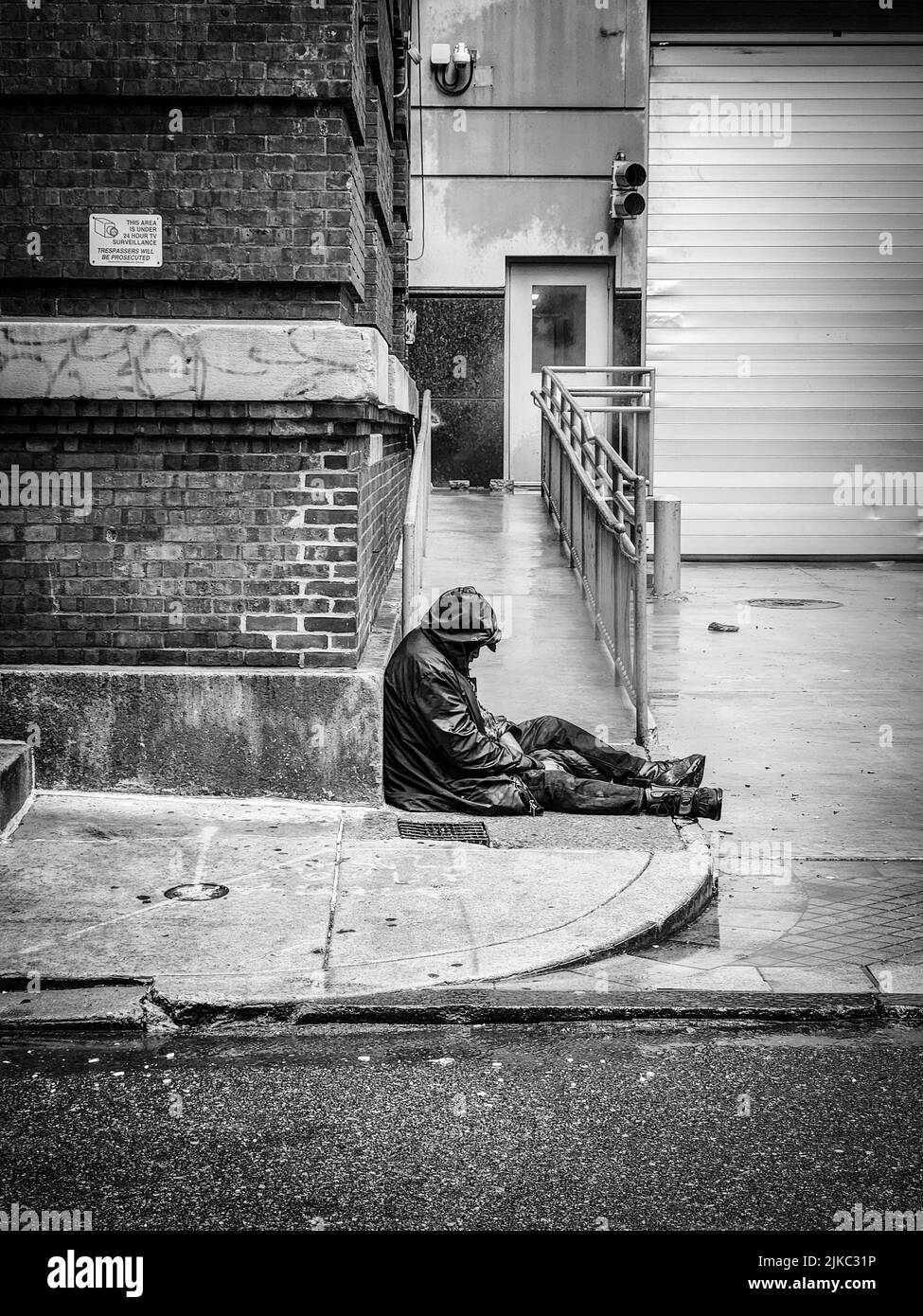 A vertical black and white shot of a homeless man sitting on the sidewalk near an apartment building Stock Photo