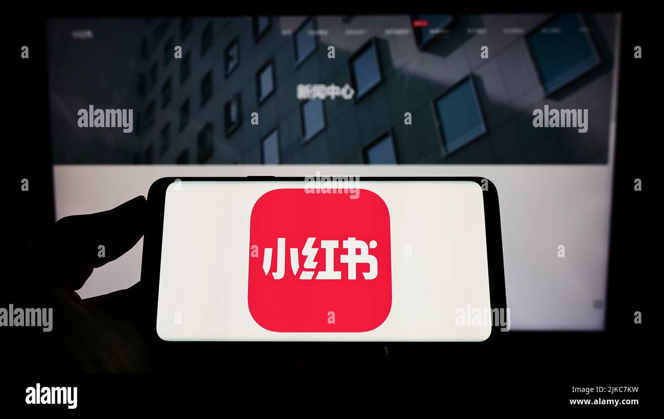 Person holding mobile phone with logo of Chinese social media company Xiaohongshu on screen in front of business web page. Focus on phone display. Stock Photo