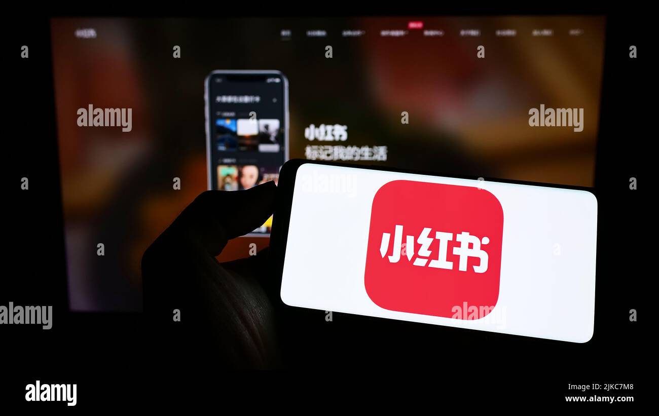 Person holding cellphone with logo of Chinese social media company Xiaohongshu on screen in front of business webpage. Focus on phone display. Stock Photo