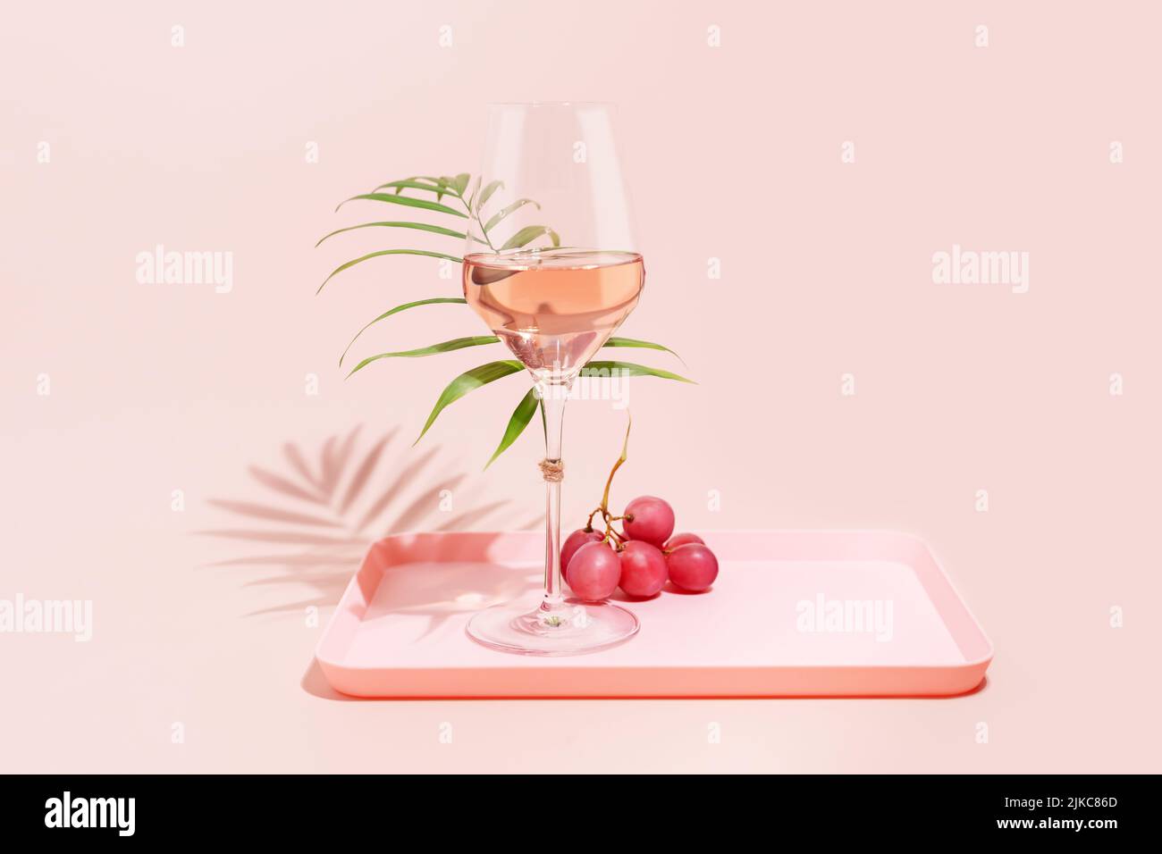 Rose wine. Minimalistic, Summer, holiday composition with glass of rose wine, grapes and palm leaf on a pink tray on a pink background. Self-partnered Stock Photo