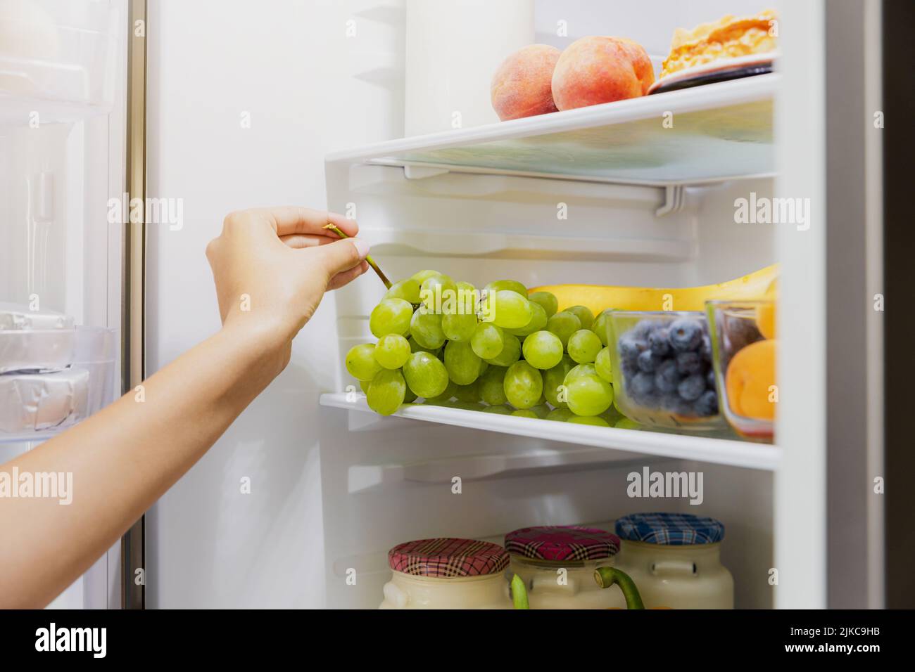 Woman hand taking, grabbing or picks up green bunch of grapes out of open refrigerator shelf or fridge drawer full of fruits, blueberries, bottles with yogurt. Healthy food diet, lifestyle concept Stock Photo
