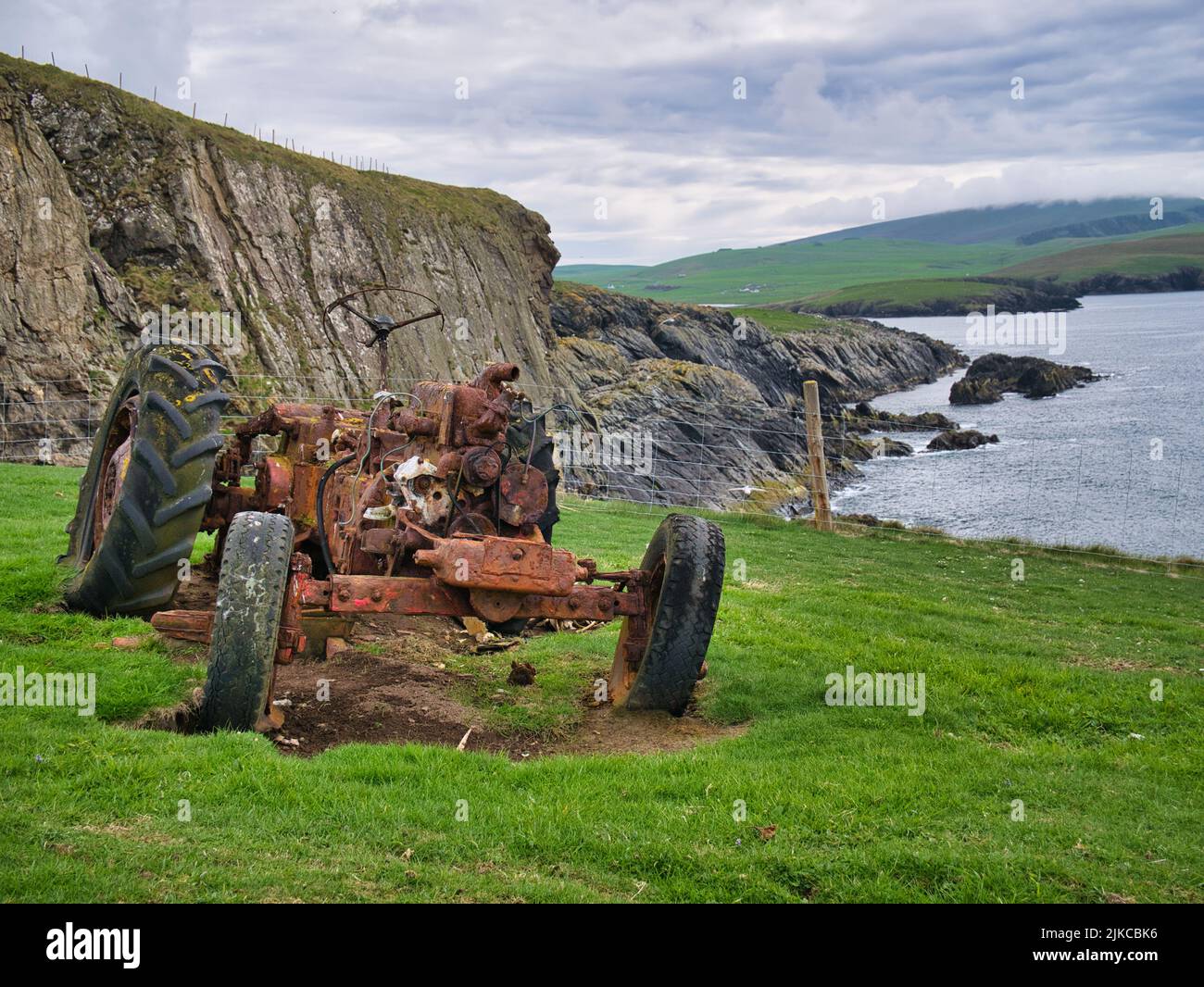 The remains of an abandoned, rusted tractor in a grassy field on the coast near Bigton, Shetland, UK. Taken on an overcast, cloudy day. Stock Photo