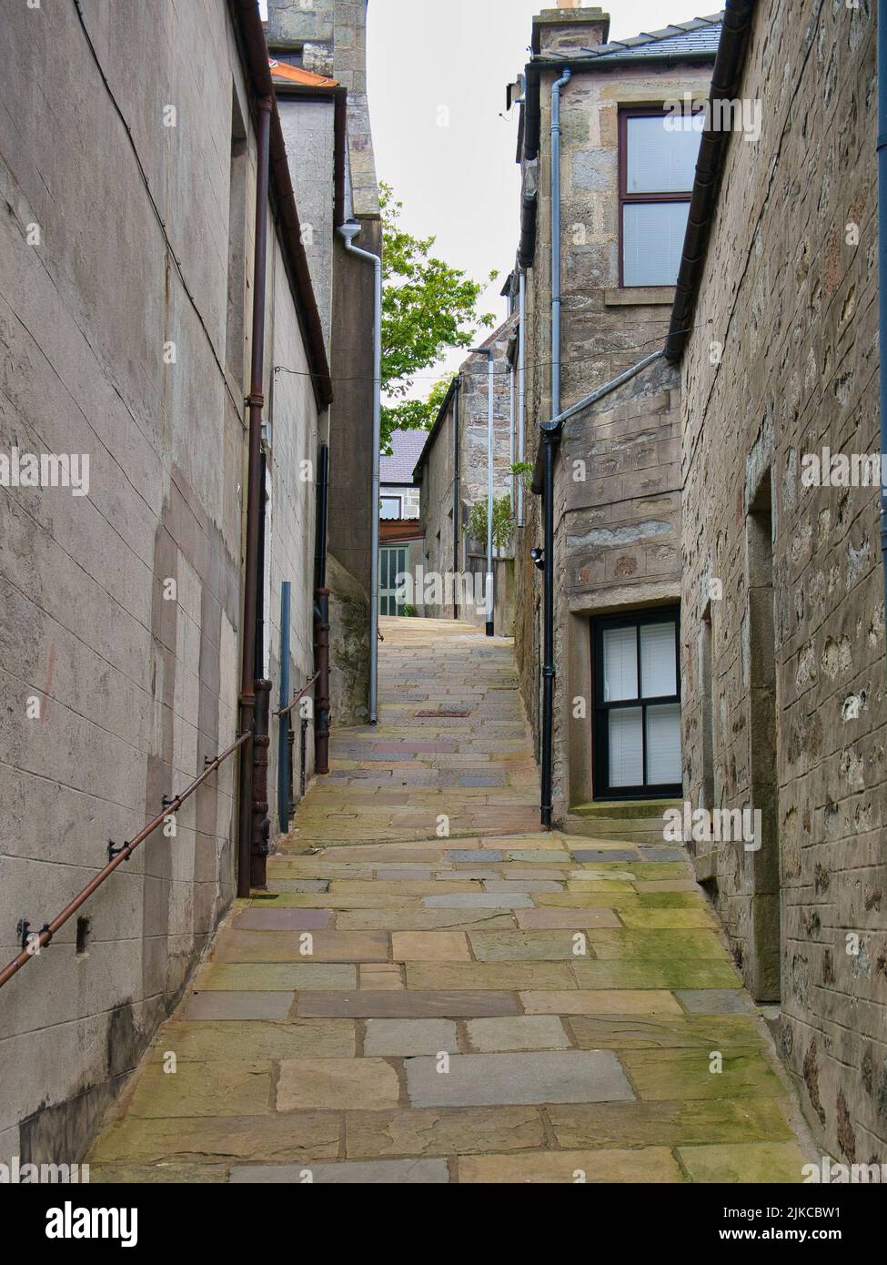 A narrow, steep footpath with green moss on some flagstones between buildings in Lerwick, Shetland, UK. A handrail appears on the left. Stock Photo
