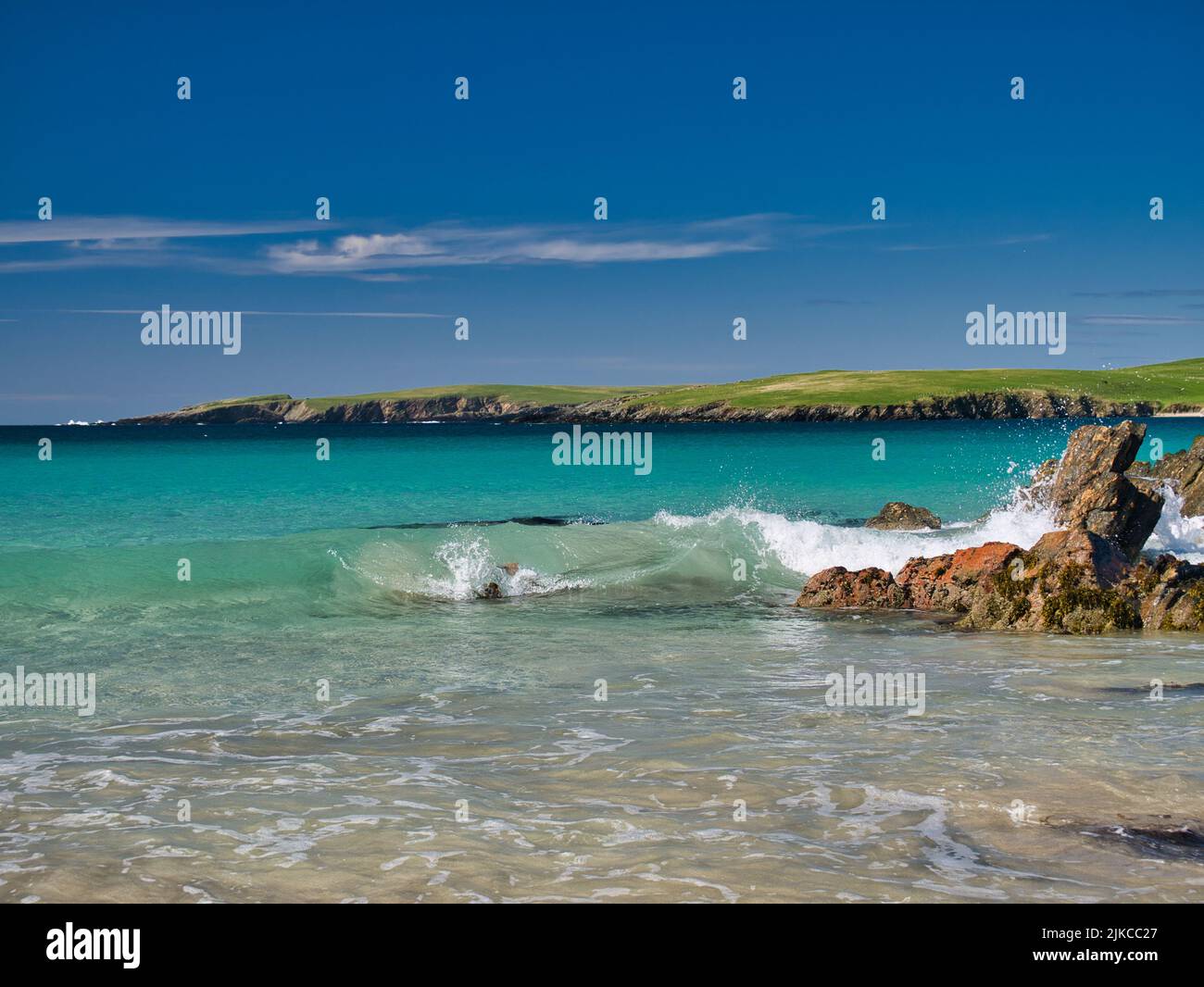 Waves break in the clear turquoise water on a beach near Scousburgh in southern Shetland, UK. Taken on a sunny day with a blue sky. Stock Photo