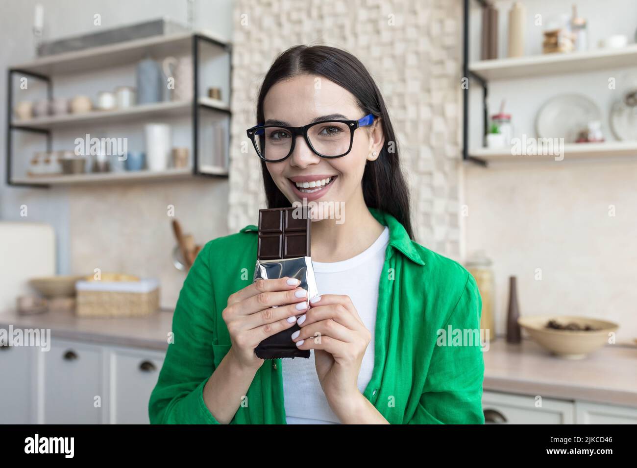 Young beautiful brunette woman in glasses and green shirt at home in kitchen, happy and smiling looking at camera eating dark chocolate Stock Photo