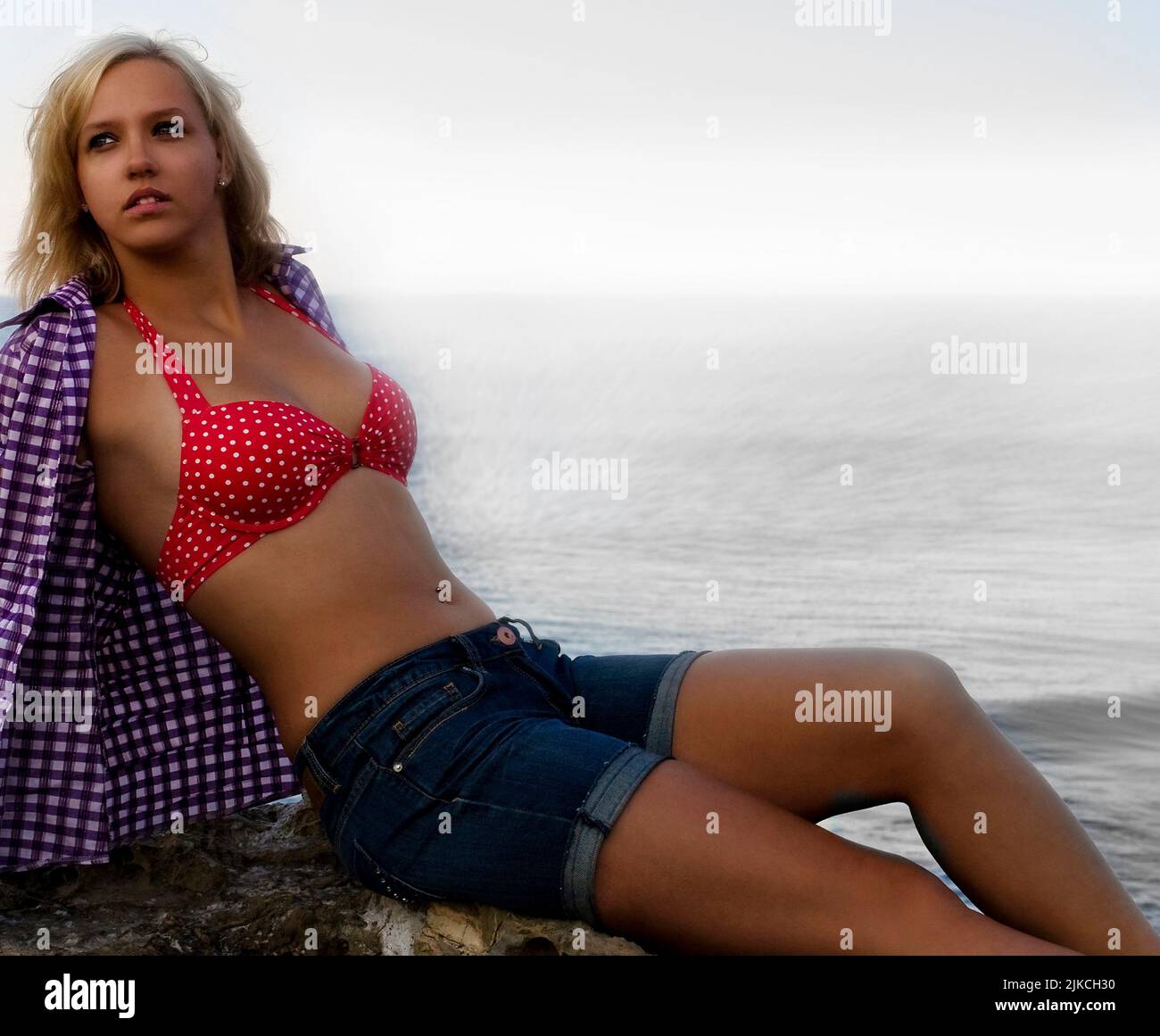 A beautiful blond Caucasian woman in a bikini top, flannel shirt and shorts, posing near a calm sea Stock Photo