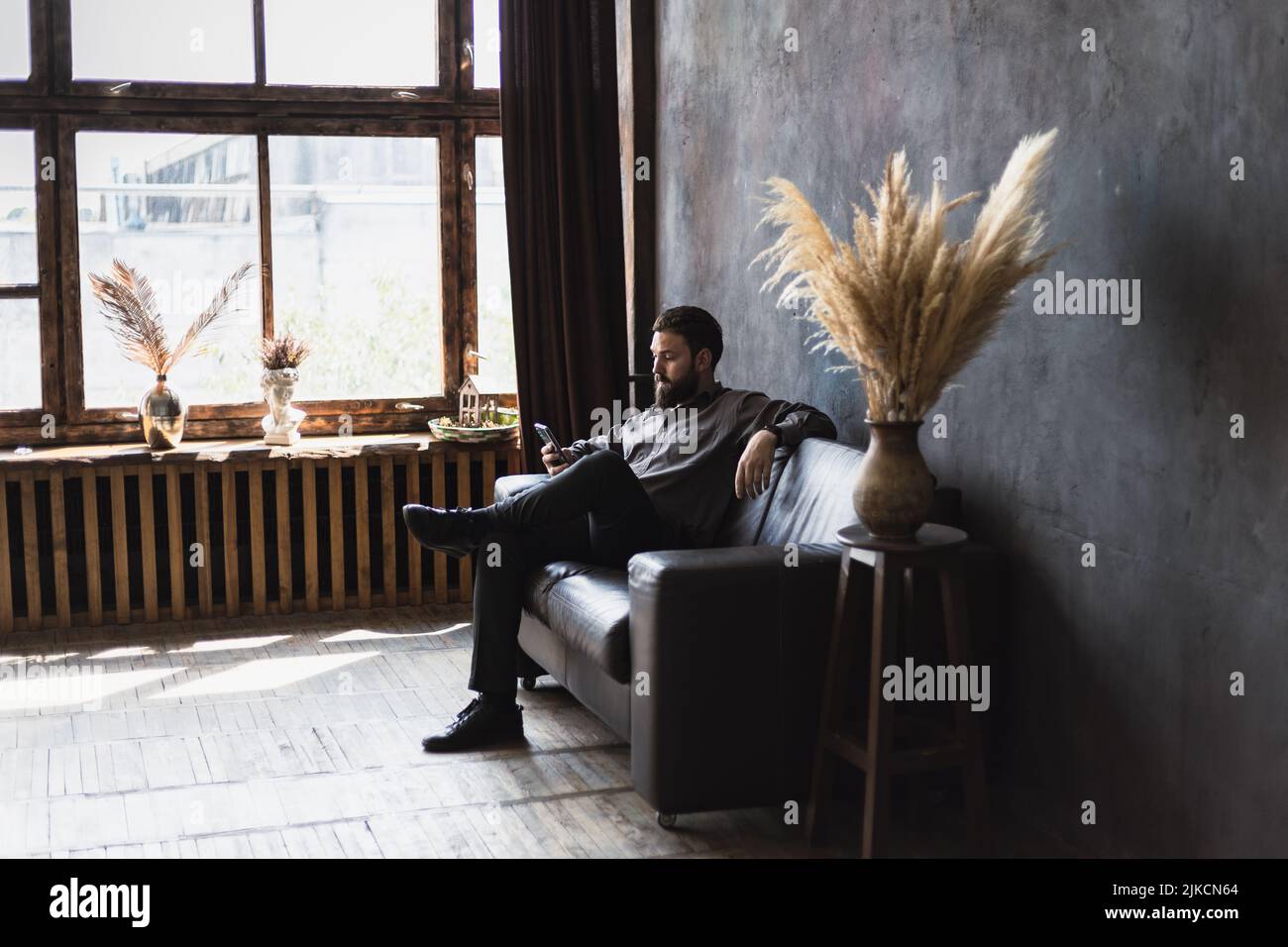 A man with a beard and a smartphone is sitting on a leather sofa. Stock Photo