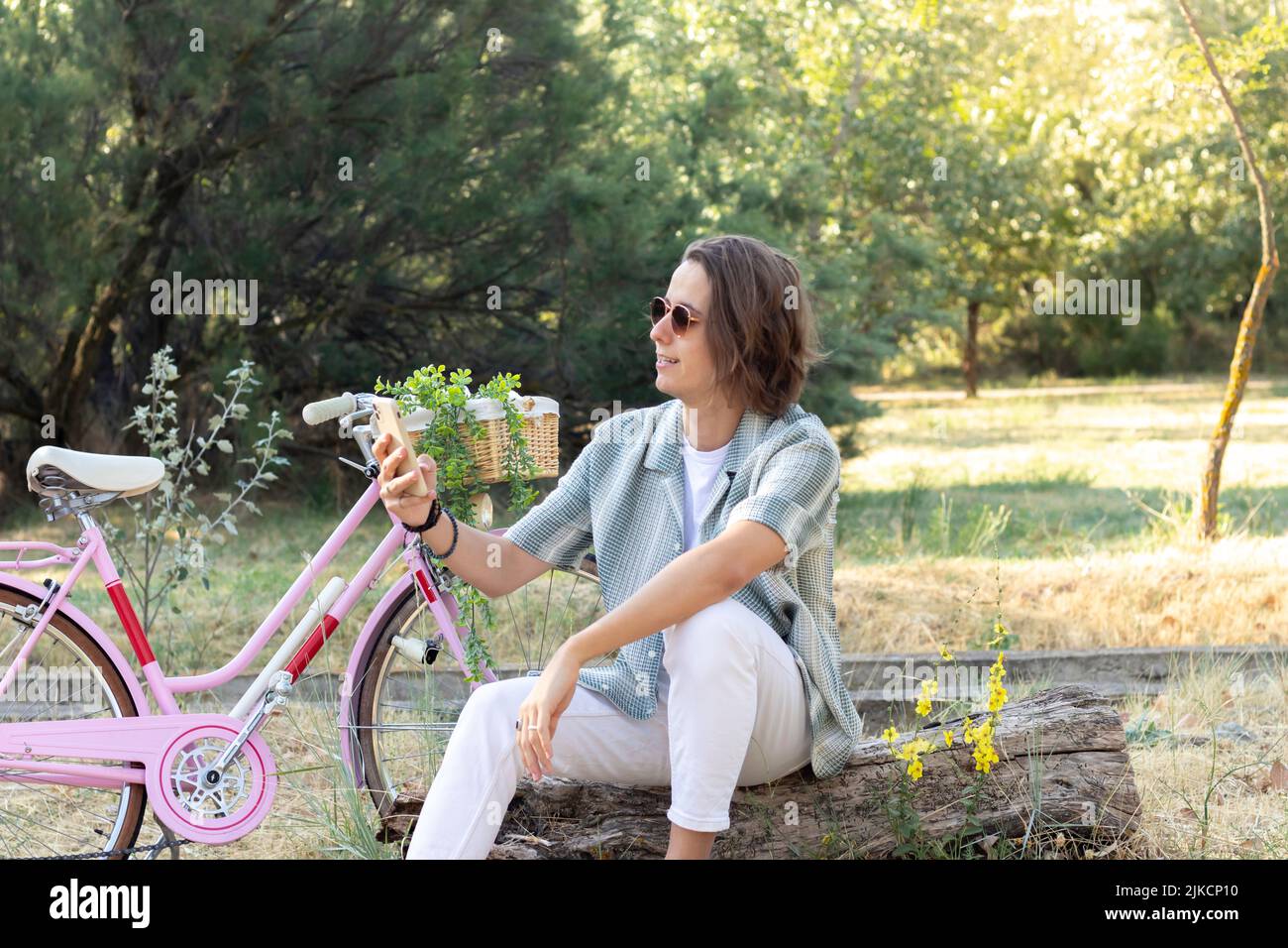 Young man sitting on a tree trunk texting his friends on the smartphone with a pink vintage bike and plants in the background Stock Photo
