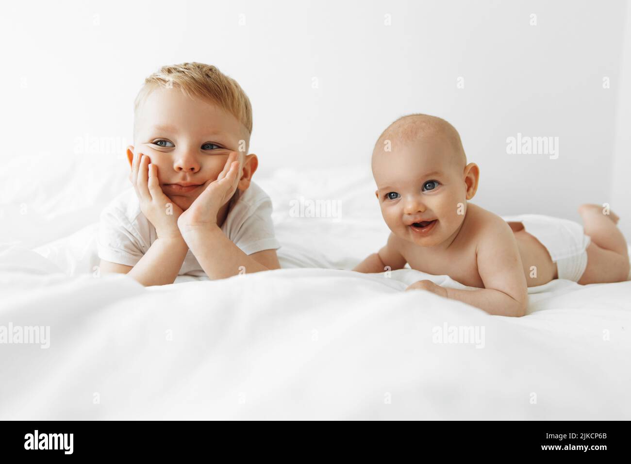 Two happy children lie on a white bed in a room. Cute older brother ...