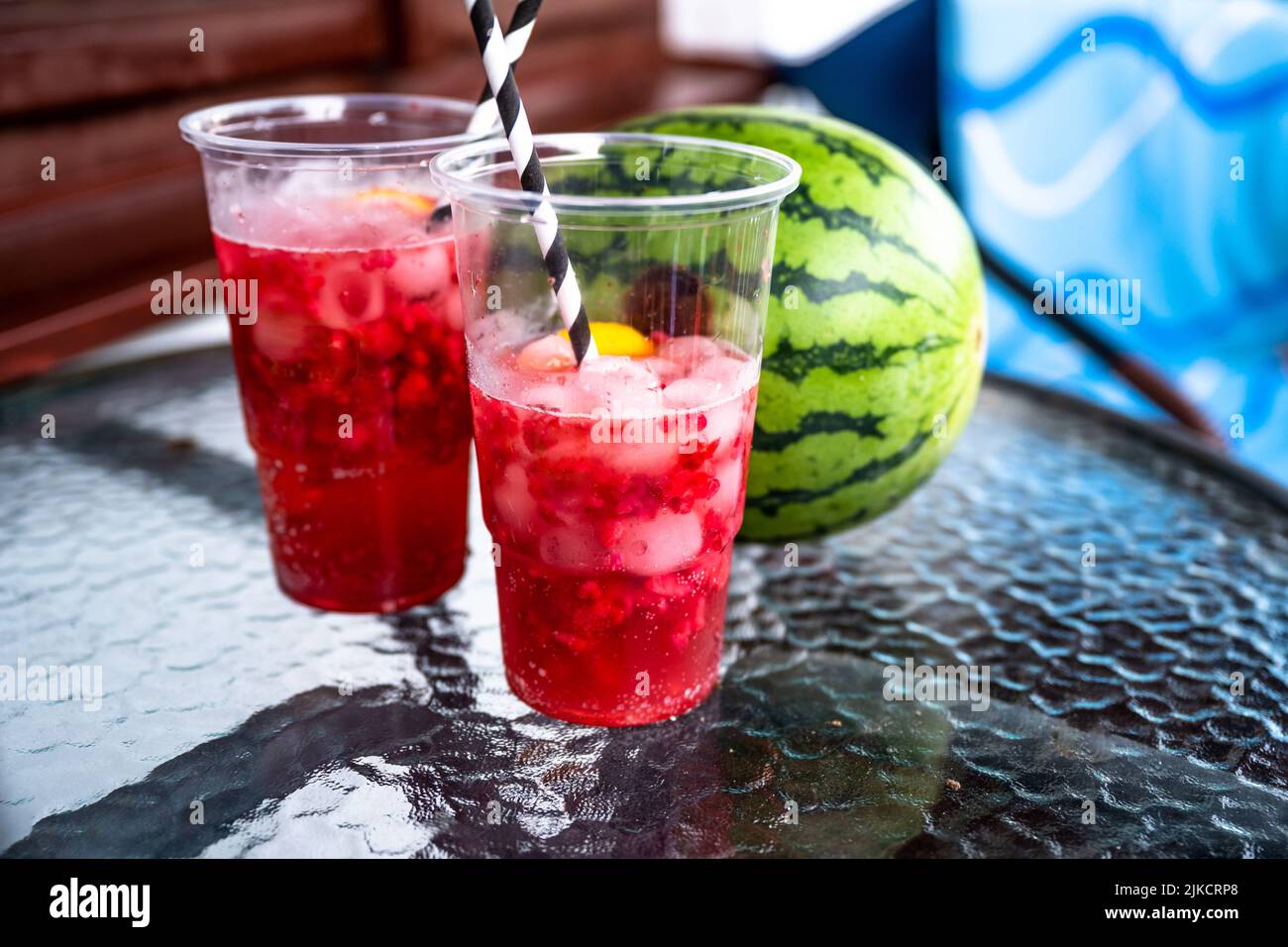 Two plastic cup with red dewy cold fruit homemade lemonade with paper straw, whole watermelone on glass table, blue and brown background. Stock Photo