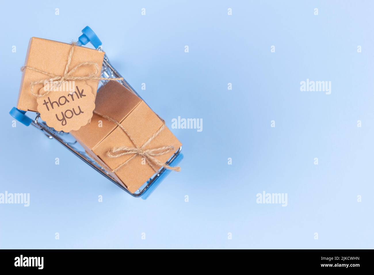 Mini shopping cart on light blue background. Kraft paper boxes with tag inscription Thank you inside trolley. Top view. E-commerce, online shopping Stock Photo