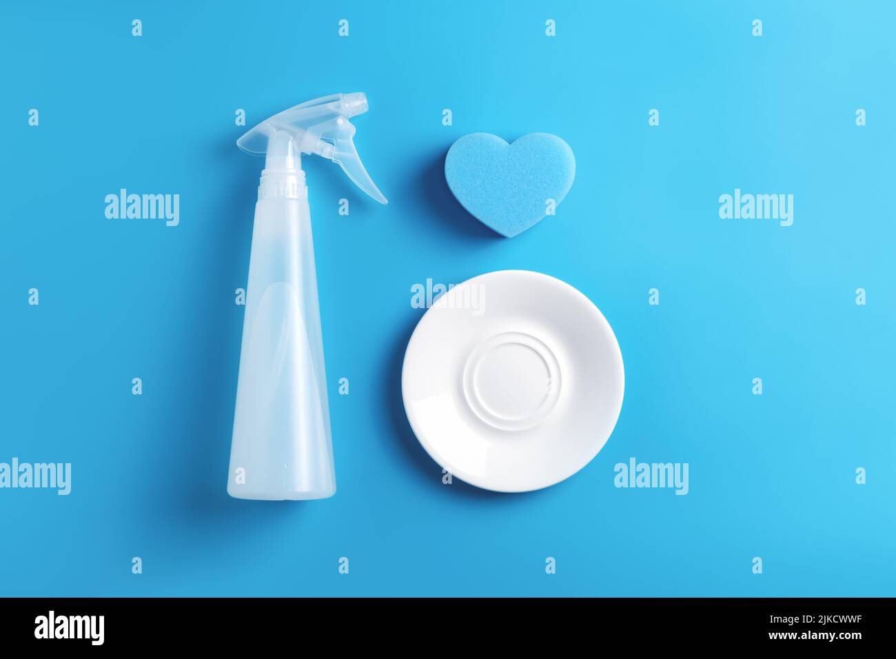 Kitchen flat lay with blue heart sponge, transparent dish cleaning spray bottle and white glass clean plate on blue background. Dishwashing concept. Stock Photo