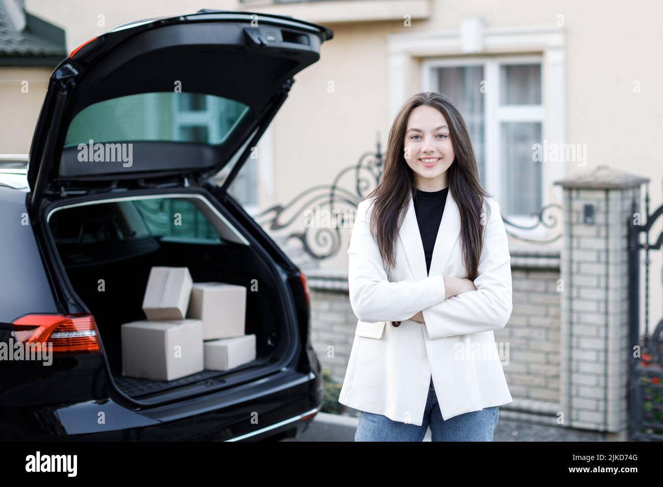 A young girl stands near the open trunk of a car, looks at the camera and smiles. Stock Photo