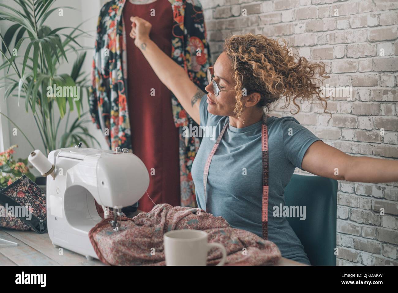 Woman outstretching arms after work on sewing machine at home or design clothes studio. Female stretching body after overwork with clothes reparation Stock Photo