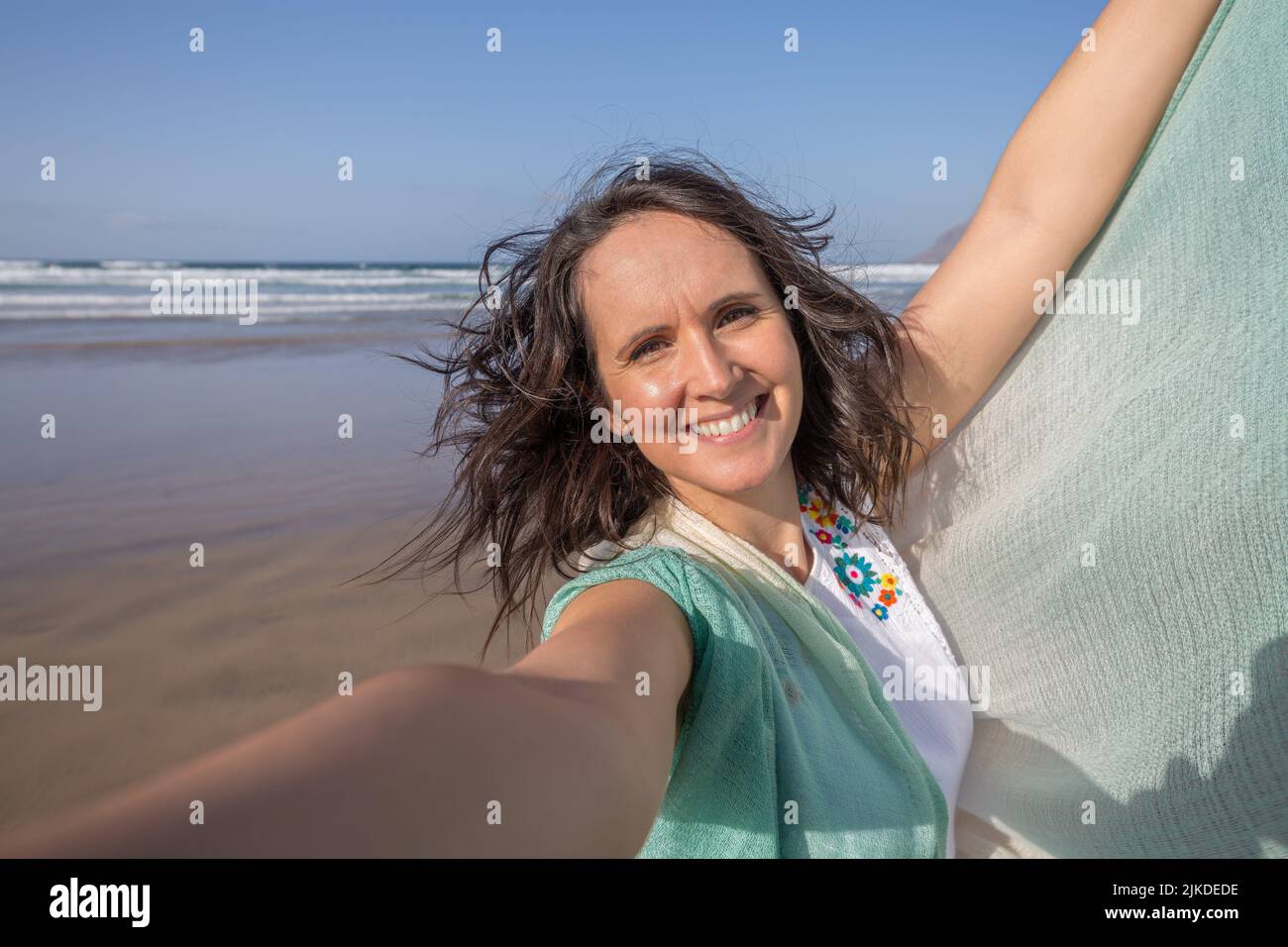 Merry middle aged female tourist with dark hair raising arm with scarf and looking at camera with smile while taking selfie against waving sea and Stock Photo