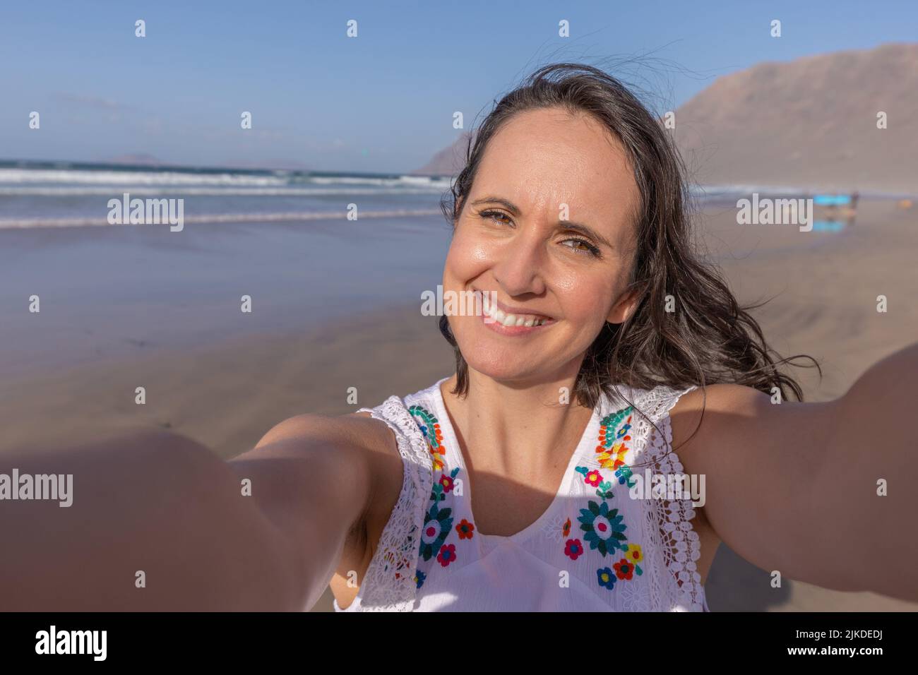 Cheerful middle aged woman with dark hair smiling and looking at camera while taking selfie on sandy beach near waving sea on summer weekend day. Stock Photo