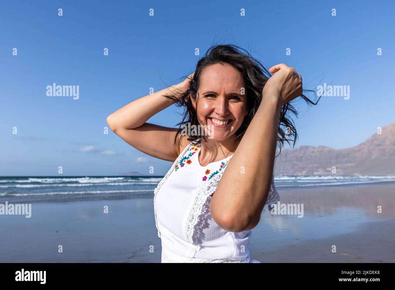 Happy middle aged brunette looking at camera with smile and touching dark hair while spending time on beach near waving sea against blue sky. Stock Photo