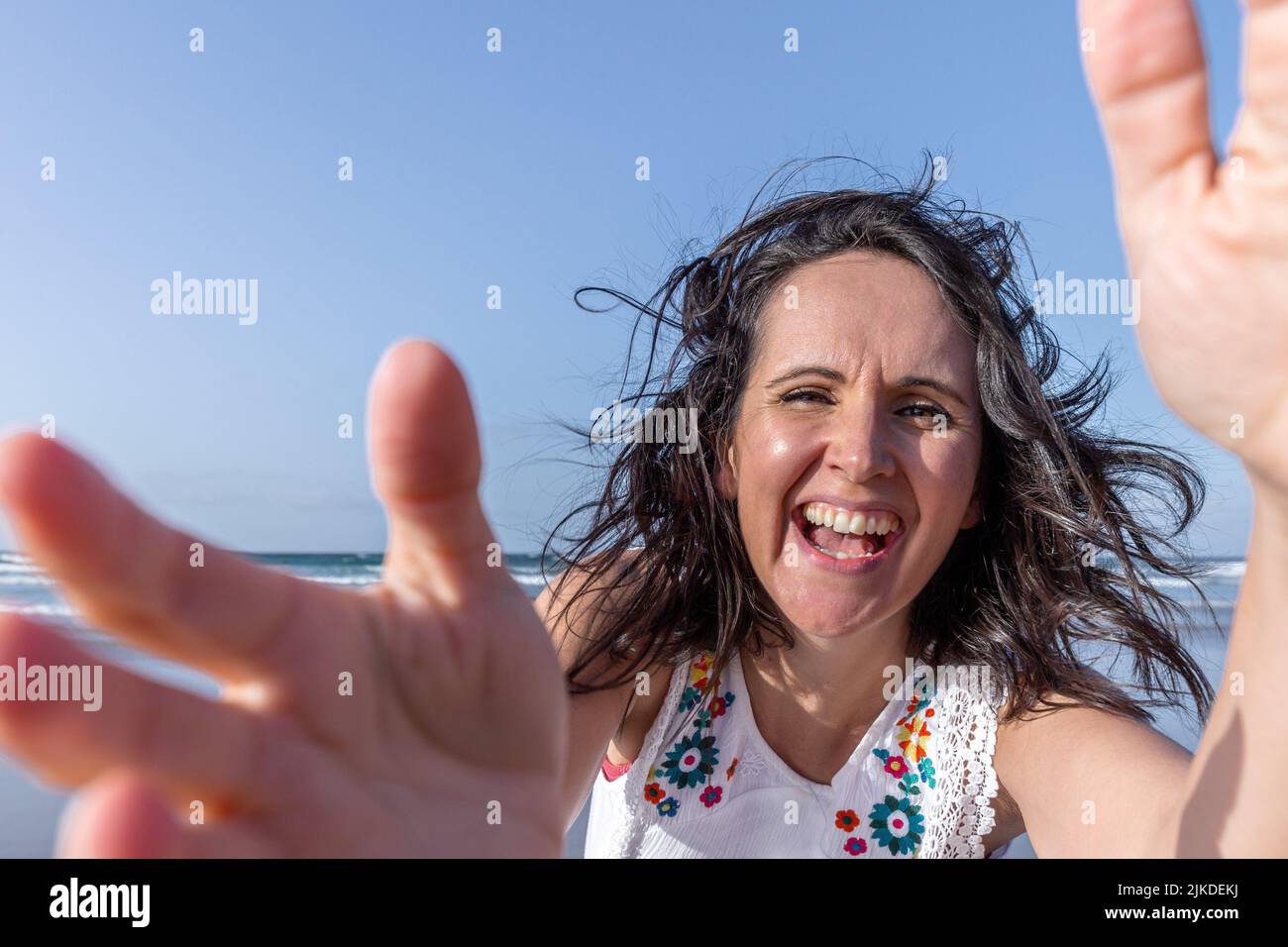 Optimistic middle aged woman with dark hair laughing and touching camera while having fun near sea against blue sky. Stock Photo