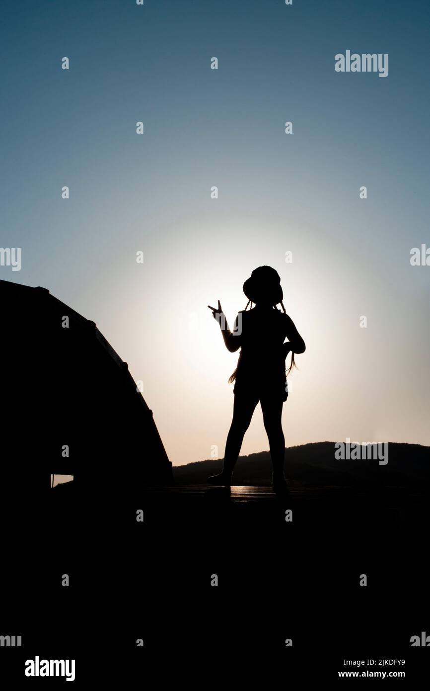 girl silhouette on a bridge holding up two fingers as a victory sign, against bright evening sun. Stock Photo