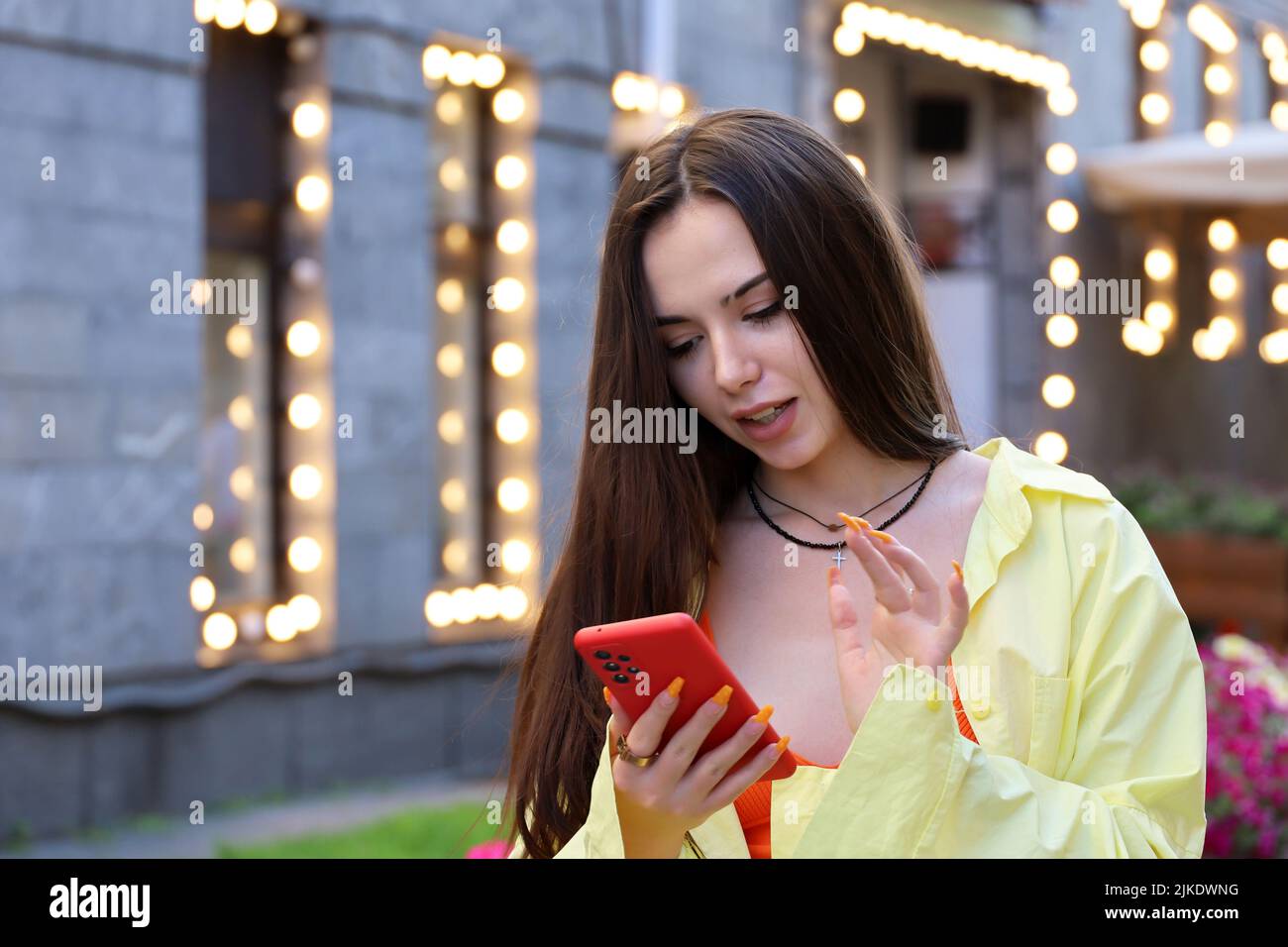 Happy attractive girl with long hair and perfect makeup using smartphone on a street on festive lights background. Female beauty, online communication Stock Photo