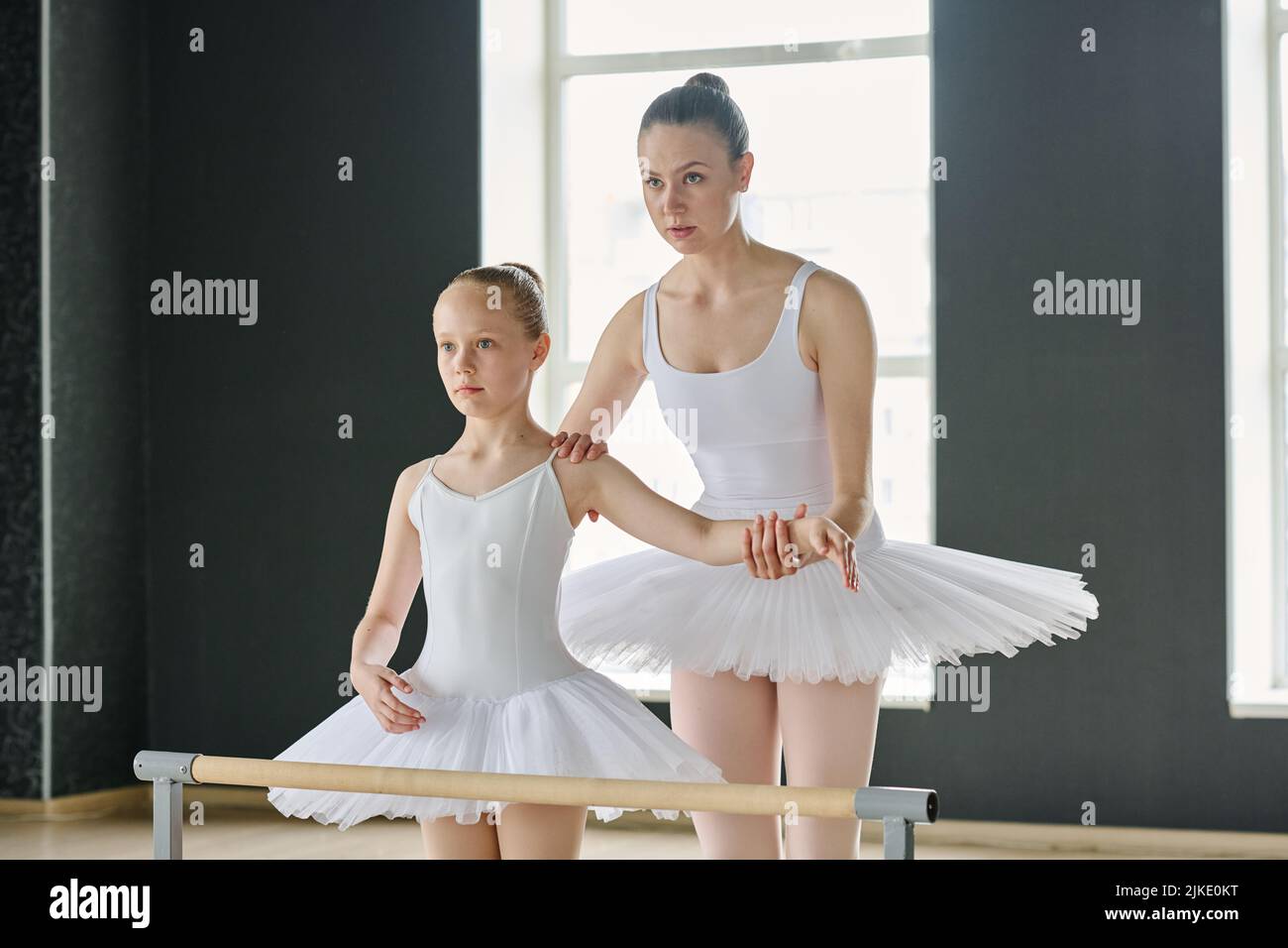 Young ballet instructor holding arm of youthful girl in white tutu while explaining her how to keep balance during performance Stock Photo