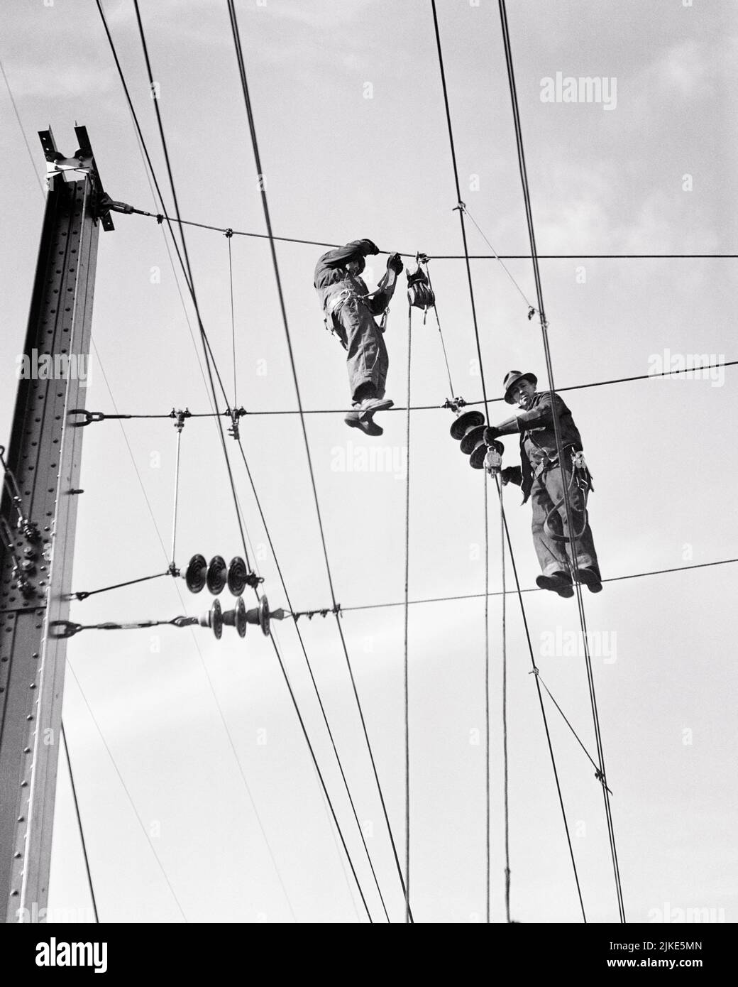1930s TWO MEN ELECTRICAL CONSTRUCTION WORKERS CLIMBING STANDING ON WIRES ELECTRIFYING RAILROAD TRACK - i1693 HAR001 HARS SKILL OCCUPATION STRUCTURE SKILLS BUILD DANGEROUS STRENGTH COURAGE BASE CAREERS KNOWLEDGE LOW ANGLE PROGRESS RECOVERY LABOR PRIDE BETTER EMPLOYMENT LINES OCCUPATIONS CONCEPTUAL INFRASTRUCTURE EMPLOYEE FACILITIES COOPERATION CORE MID-ADULT MID-ADULT MAN PRECISION SYSTEMS BLACK AND WHITE CAUCASIAN ETHNICITY ECONOMY HAR001 LABORING OLD FASHIONED Stock Photo