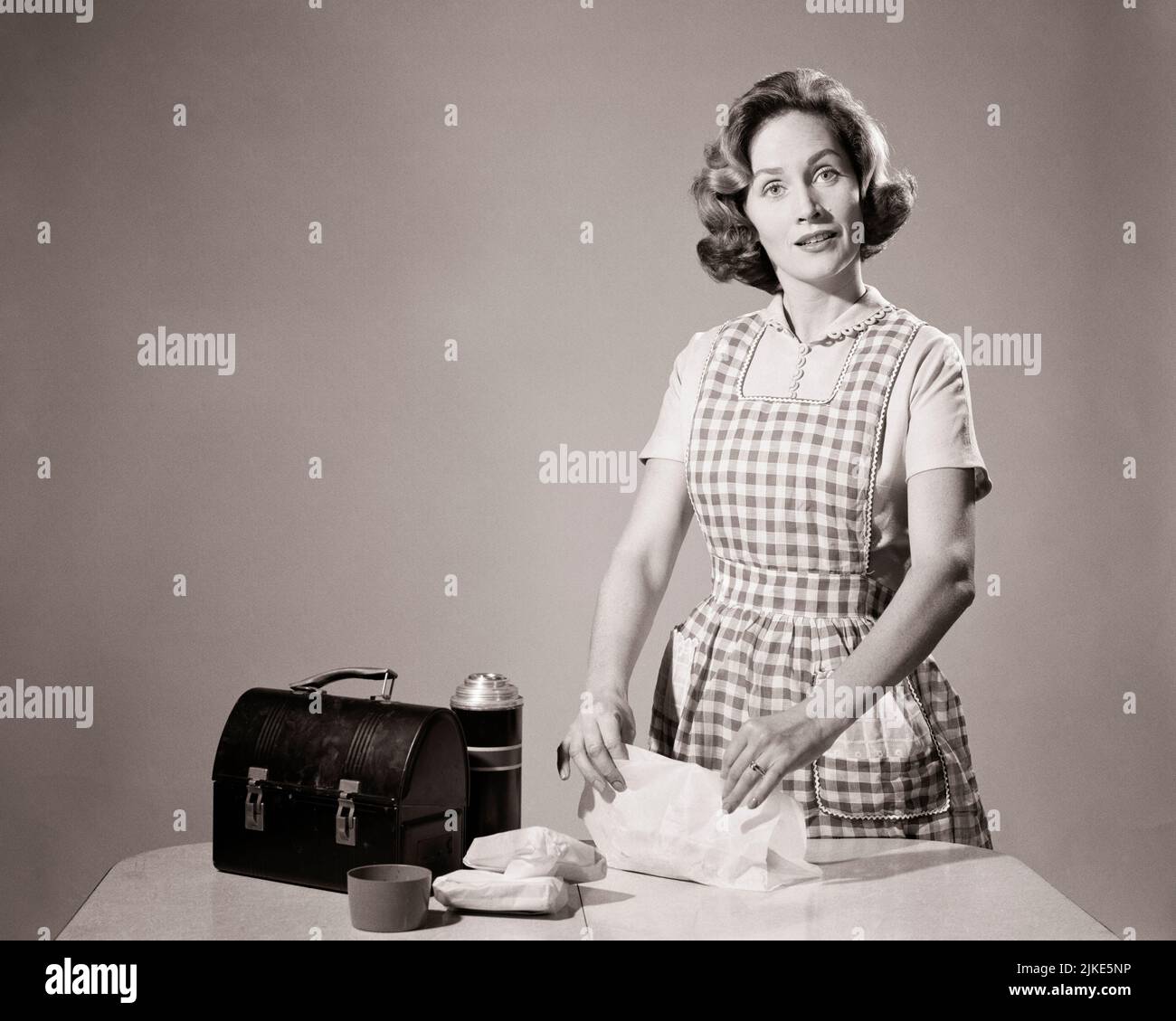 1960s WOMAN HOUSEWIFE WEARING CHECKERED APRON LOOKING AT CAMERA PREPARING PACKING LUNCH BOX AND THERMOS BOTTLE IN KITCHEN - h3321 DEB001 HARS CARING B&W EYE CONTACT HOMEMAKER HOMEMAKERS CHORE AND THERMOS HOUSEWIVES TASKS DEB001 CHECKERED MID-ADULT MID-ADULT WOMAN TASK UNFULFILLED WIVES YOUNG ADULT WOMAN BLACK AND WHITE CAUCASIAN ETHNICITY OLD FASHIONED Stock Photo