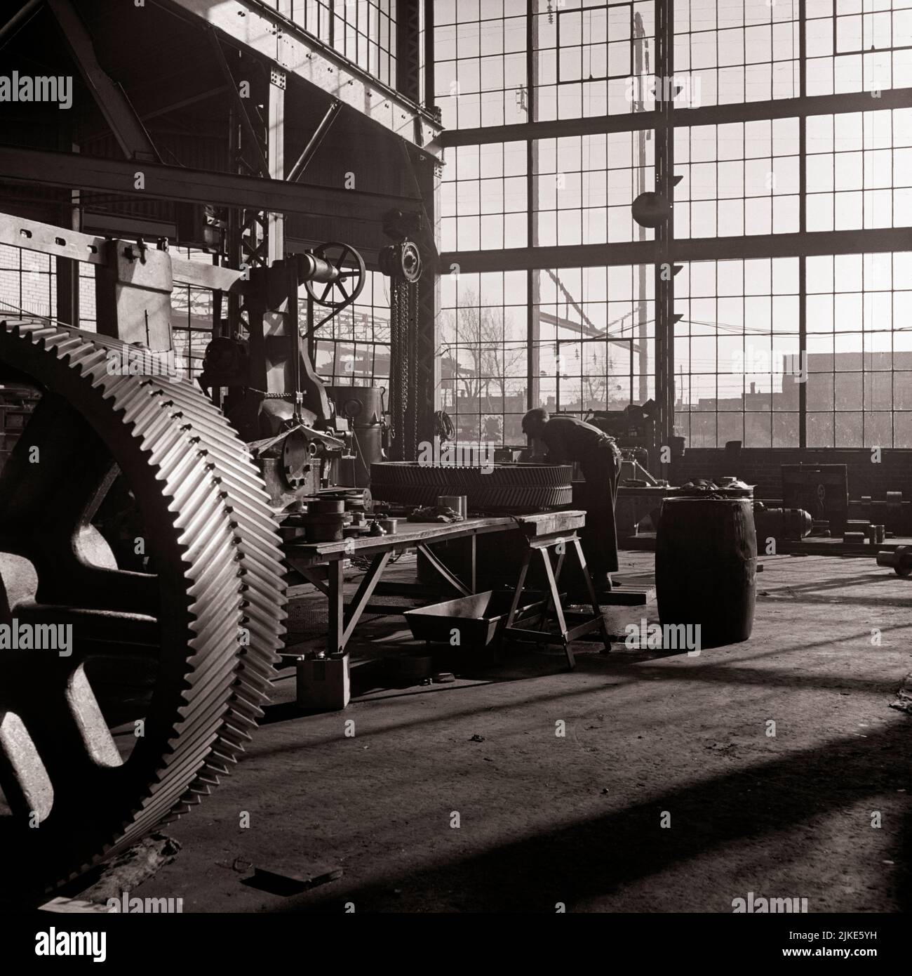 1930s SINGLE ANONYMOUS SILHOUETTED MACHINIST WORKING IN GLASS WALLED FACTORY MANUFACTURING LARGE INDUSTRIAL GEARS - i3446 HAR001 HARS OCCUPATIONS CONCEPTUAL INFRASTRUCTURE ANONYMOUS EMPLOYEE FACILITIES CORE SYSTEMS BLACK AND WHITE ECONOMY HAR001 LABORING OLD FASHIONED Stock Photo