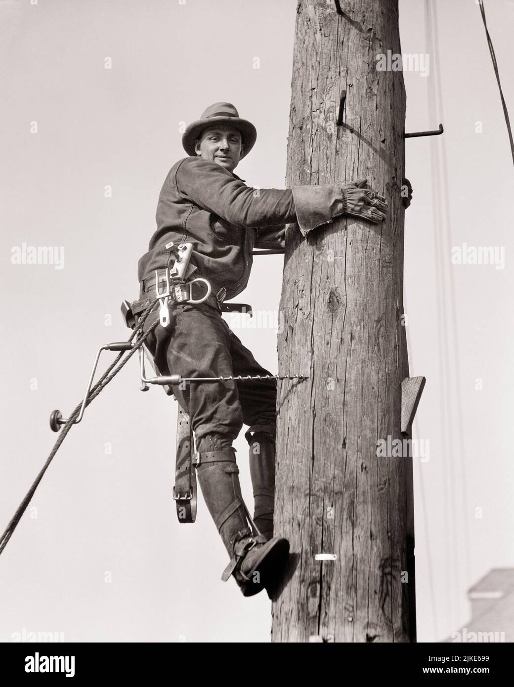 1930s ONE MAN WORKING ON UTILITY POLE SMILING LOOKING AT CAMERA - i606 HAR001 HARS OCCUPATION STRUCTURE SKILLS CHEERFUL BUILD BASE CAREERS LOW ANGLE RECOVERY LABOR BETTER EMPLOYMENT OCCUPATIONS SMILES JOYFUL INFRASTRUCTURE EMPLOYEE FACILITIES CORE SYSTEMS YOUNG ADULT MAN BLACK AND WHITE CAUCASIAN ETHNICITY ECONOMY HAR001 LABORING OLD FASHIONED Stock Photo