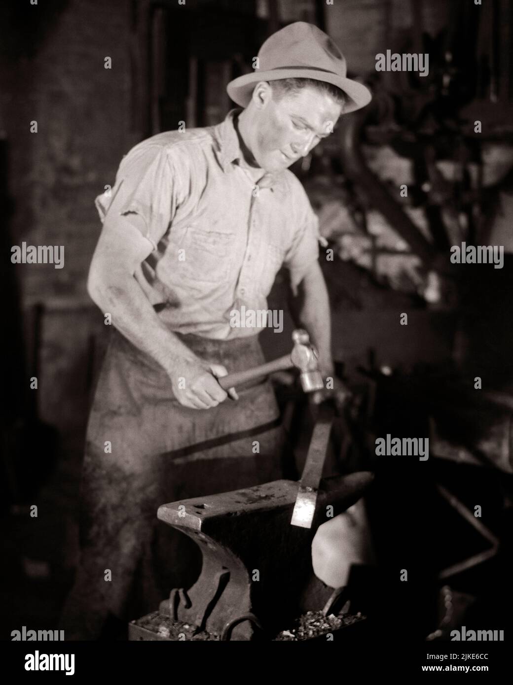 1930s MAN IN FORGE WORKING A PIECE OF HEATED METAL WITH BALL PEEN HAMMER ON AN ANVIL - i840 HAR001 HARS STRENGTH CAREERS LABOR EMPLOYMENT OCCUPATIONS CONCEPTUAL INFRASTRUCTURE EMPLOYEE HEATED ANVIL YOUNG ADULT MAN BLACK AND WHITE CAUCASIAN ETHNICITY HAR001 LABORING OLD FASHIONED Stock Photo