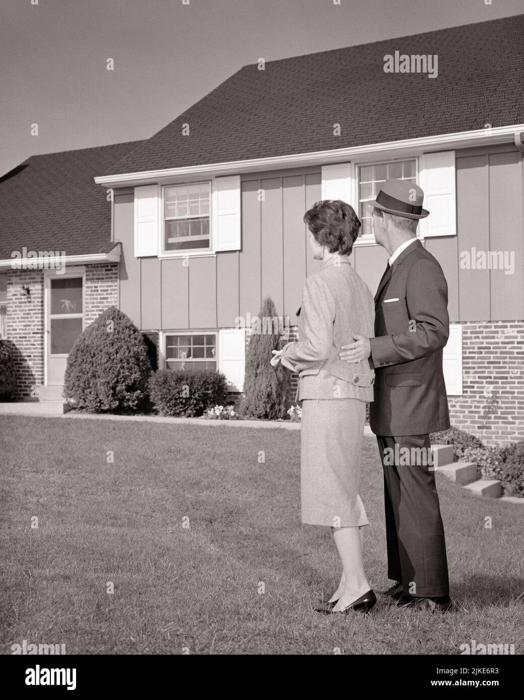 1960s BACK VIEW MAN AND WOMAN COUPLE STANDING IN FRONT LAWN LOOKING AT SPLIT LEVEL SUBURBAN HOUSE - j12435 HAR001 HARS HUSBANDS HOME LIFE COPY SPACE FULL-LENGTH LADIES PERSONS RESIDENTIAL MALES BUILDINGS B&W PARTNER SPLIT SUIT AND TIE DREAMS STRUCTURE PROPERTY AND CHOICE EXTERIOR SPLIT LEVEL REAR VIEW HOMES REAL ESTATE CONNECTION CONCEPTUAL FROM BEHIND STRUCTURES RESIDENCE STYLISH EDIFICE IN FRONT OF BACK VIEW COOPERATION LEVEL MID-ADULT MID-ADULT MAN MID-ADULT WOMAN WIVES BLACK AND WHITE CAUCASIAN ETHNICITY HAR001 OLD FASHIONED SHUTTERS Stock Photo