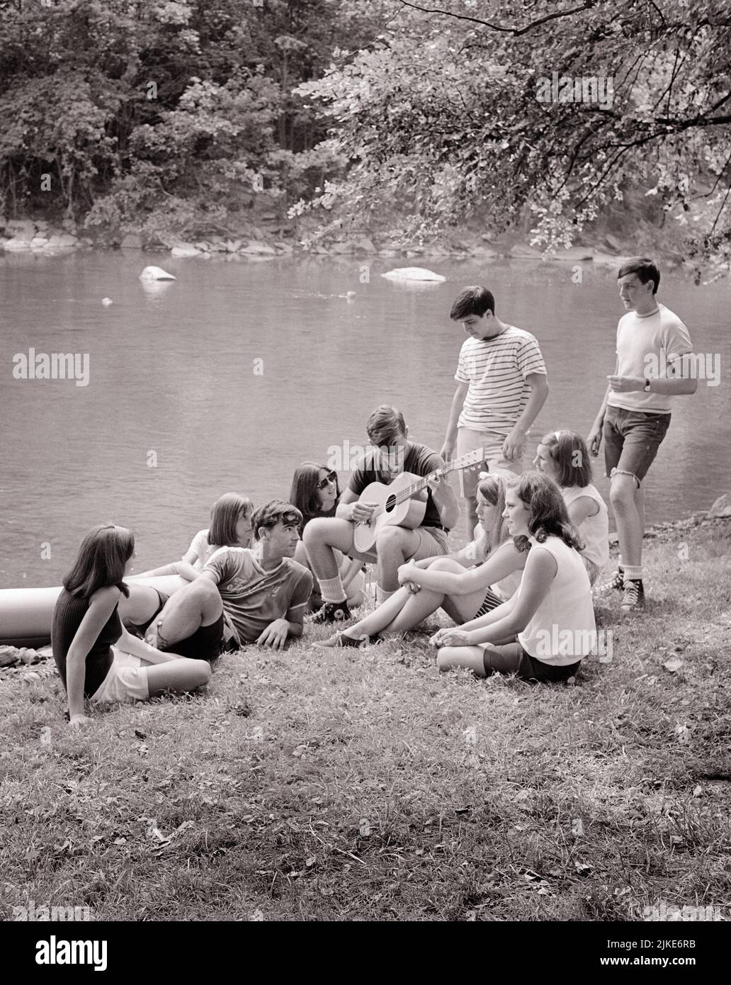 1960s GROUP OF 10 TEENAGERS SITTING TOGETHER AROUND LAKESIDE LISTENING TO ONE BOY PLAY THE GUITAR - j12363 HAR001 HARS TEENAGE GIRL TEENAGE BOY ENTERTAINMENT SIBLINGS SISTERS B&W LAKESIDE HIGH ANGLE ADVENTURE LEISURE RECREATION SIBLING CONNECTION 10 SUPPORT TEENAGED COOPERATION JUVENILES TOGETHERNESS BLACK AND WHITE CAUCASIAN ETHNICITY HAR001 OLD FASHIONED Stock Photo