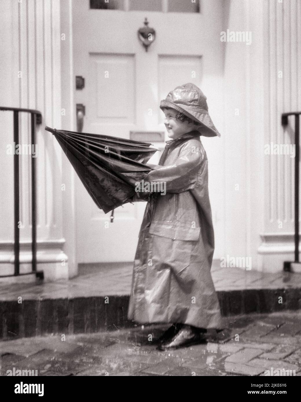 1920s LITTLE GIRL IN THE RAIN WEARING LONG RAIN COAT AND HAT SOU’WESTER FOUL WEATHER GEAR AND PUTTING UP AN UMBRELLA - j1280 HAR001 HARS WINNING HOME LIFE FULL-LENGTH RAINY RESIDENTIAL CUTOUT RAINING BUILDINGS B&W CHEERFUL ADVENTURE AND EXCITEMENT UP HOMES SMILES JOYFUL RESIDENCE FRONT DOOR FOUL PLEASANT AGREEABLE CHARMING FOUL WEATHER JUVENILES LOVABLE PLEASING ADORABLE APPEALING BLACK AND WHITE CAUCASIAN ETHNICITY HAR001 OLD FASHIONED Stock Photo