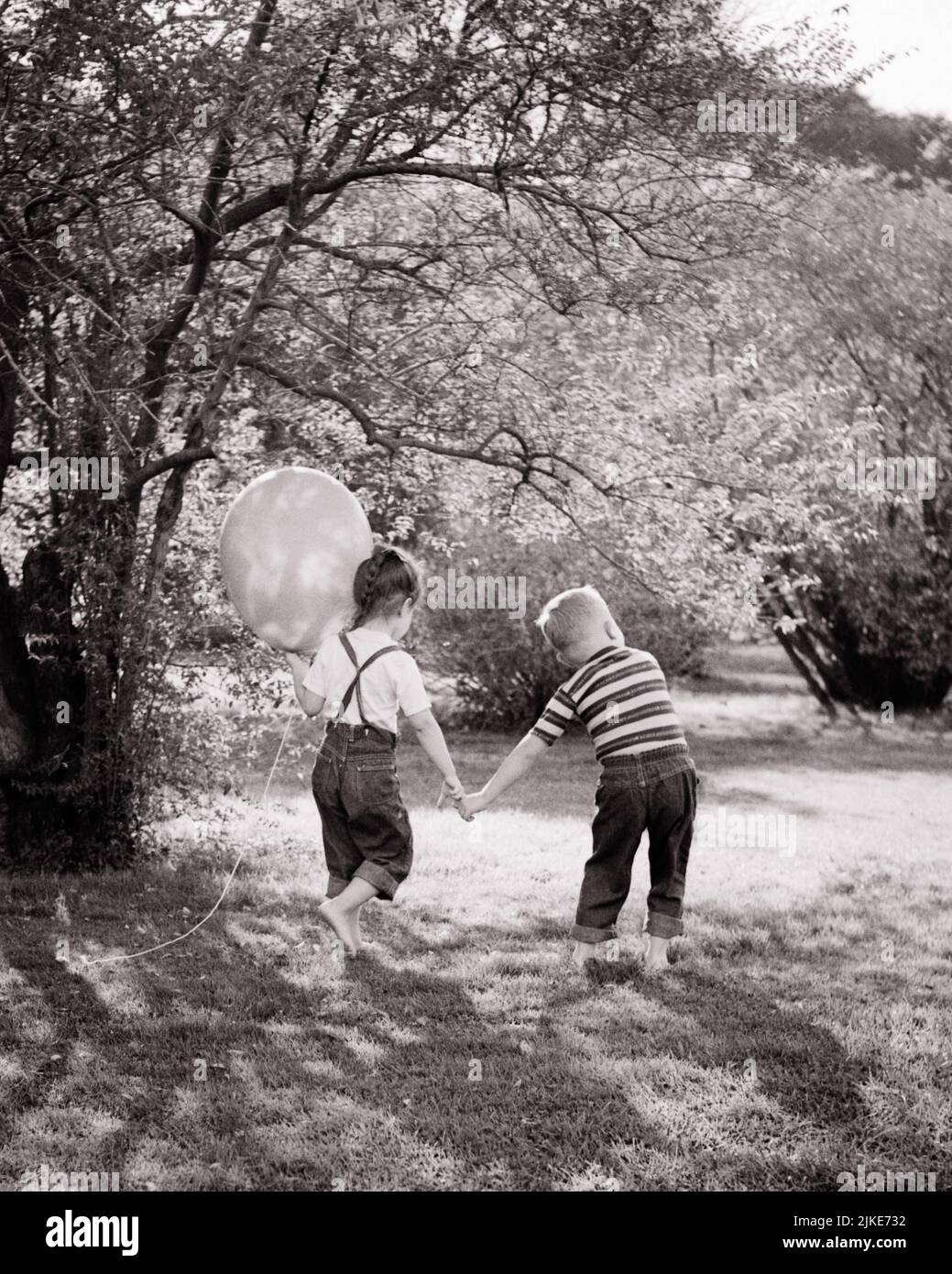 1960s LITTLE BOY AND GIRL HOLDING HANDS WALKING IN GRASS GIRL BAREFOOT HOLDING A BALLOON BOTH WEARING BLUE JEANS TEE SHIRTS - j13216 HAR001 HARS JOY LIFESTYLE FEMALES BROTHERS NATURE TEE COPY SPACE FRIENDSHIP FULL-LENGTH PERSONS MALES SIBLINGS DENIM SISTERS B&W SUMMERTIME HIGH ANGLE ADVENTURE AND SHIRTS BAREFOOT SIBLING CONNECTION FRIENDLY BLUE JEANS COOPERATION GROWTH INFORMAL JUVENILES SEASON TOGETHERNESS TWILL BLACK AND WHITE CASUAL CAUCASIAN ETHNICITY HAR001 OLD FASHIONED Stock Photo