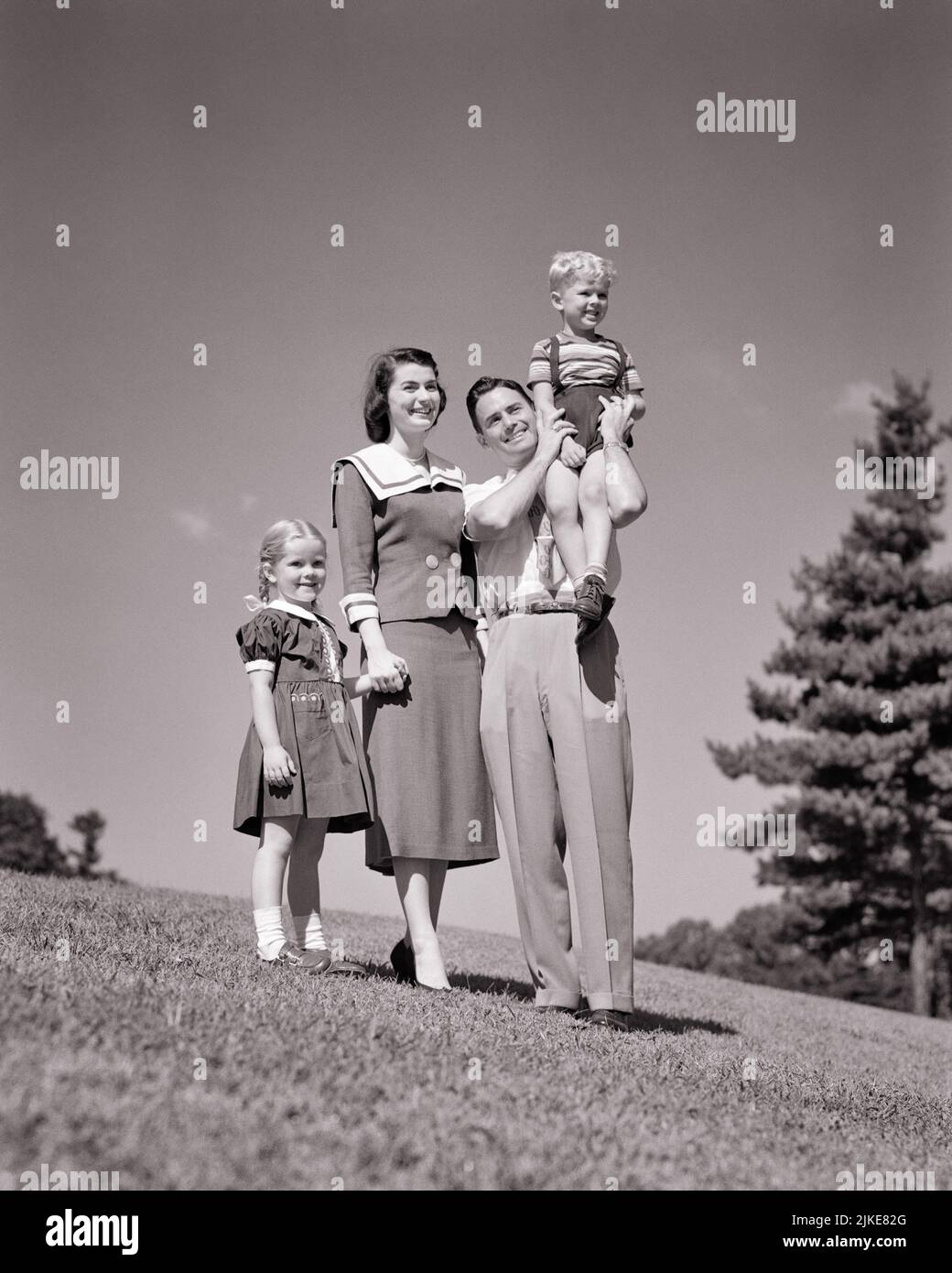 1950s FAMILY ON GRASSY HILLSIDE MOTHER FATHER AND 2 KIDS BOY ON DAD’S SHOULDER GIRL HOLDING HANDS WITH MOM LOOKING OUT AT VIEW - j4275 HAR001 HARS SHOULDER MOM CLOTHING NOSTALGIC PAIR 4 SUBURBAN MOTHERS OLD TIME NOSTALGIA BROTHER OLD FASHION SISTER JUVENILE STYLE PLEASED FAMILIES JOY LIFESTYLE FEMALES MARRIED BROTHERS RURAL SPOUSE HUSBANDS HEALTHINESS HOME LIFE COPY SPACE FRIENDSHIP FULL-LENGTH LADIES PERSONS MALES SIBLINGS SISTERS FATHERS B&W PARTNER SUMMERTIME HAPPINESS CHEERFUL ADVENTURE LEISURE AND DADS EXCITEMENT LOW ANGLE PARKS RECREATION PRIDE SIBLING SMILES CONNECTION NUCLEAR FAMILY Stock Photo