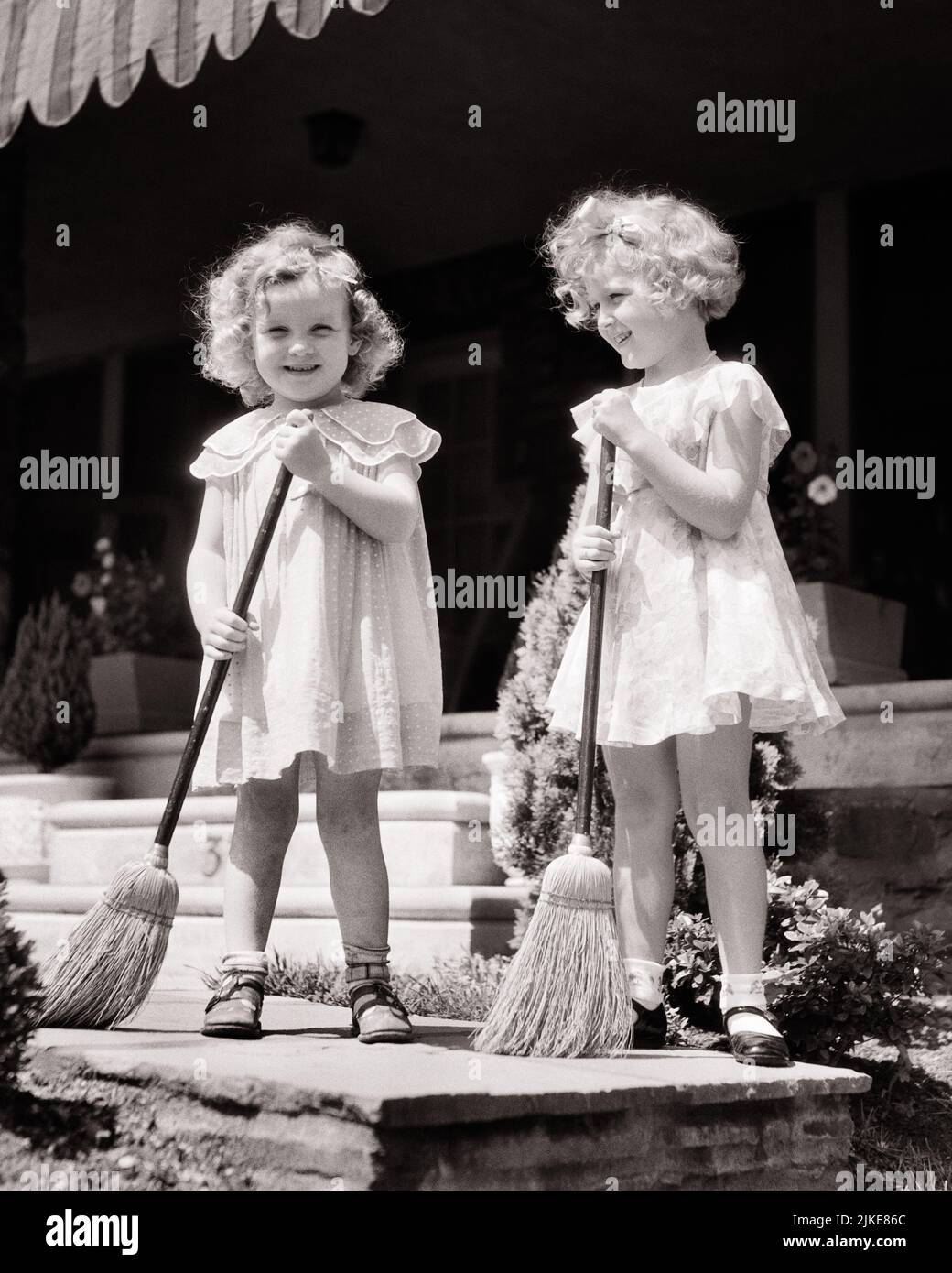 1930s TWO SMILING CURLY HAIRED GIRLS IN CUTE SUMMER DRESSES HOLDING BROOMS SWEEPING THE SIDEWALK IN FRONT OF THE HOUSE PORCH - j5366 HAR001 HARS FRIEND TEAMWORK TWIN IDENTICAL DOUBLE PLEASED JOY LIFESTYLE SATISFACTION FEMALES HOUSES HEALTHINESS HOME LIFE COPY SPACE FRIENDSHIP FULL-LENGTH BROOM RESIDENTIAL MATCH BUILDINGS SIBLINGS SISTERS B&W SUMMERTIME EYE CONTACT DRESSES GOALS MATCHING SAME HAPPINESS CHEERFUL SWEEPING LOW ANGLE CURLY MARY JANE PRIDE BROOMS HAIRED HOMES SIBLING SMILES FRIENDLY JOYFUL RESIDENCE STYLISH SUPPORT IN FRONT OF LOOK-ALIKE COOPERATION DUPLICATE GROWTH JUVENILES Stock Photo
