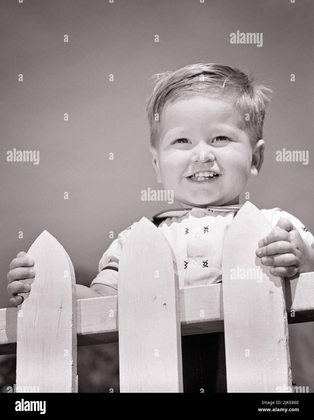 1950s PORTRAIT TODDLER BOY SMILING STANDING AT WHITE PICKET FENCE LOOKING AT CAMERA - j5411 HAR001 HARS HEALTHINESS HOME LIFE COPY SPACE MALES CONFIDENCE B&W EYE CONTACT HAPPINESS HEAD AND SHOULDERS CHEERFUL LEADERSHIP LOW ANGLE HANDSOME PRIDE SMILES JOYFUL BABY BOY PLEASANT AGREEABLE CHARMING EAGER GROWTH JUVENILES LOVABLE PLEASING ADORABLE APPEALING BLACK AND WHITE CAUCASIAN ETHNICITY HAR001 OLD FASHIONED Stock Photo