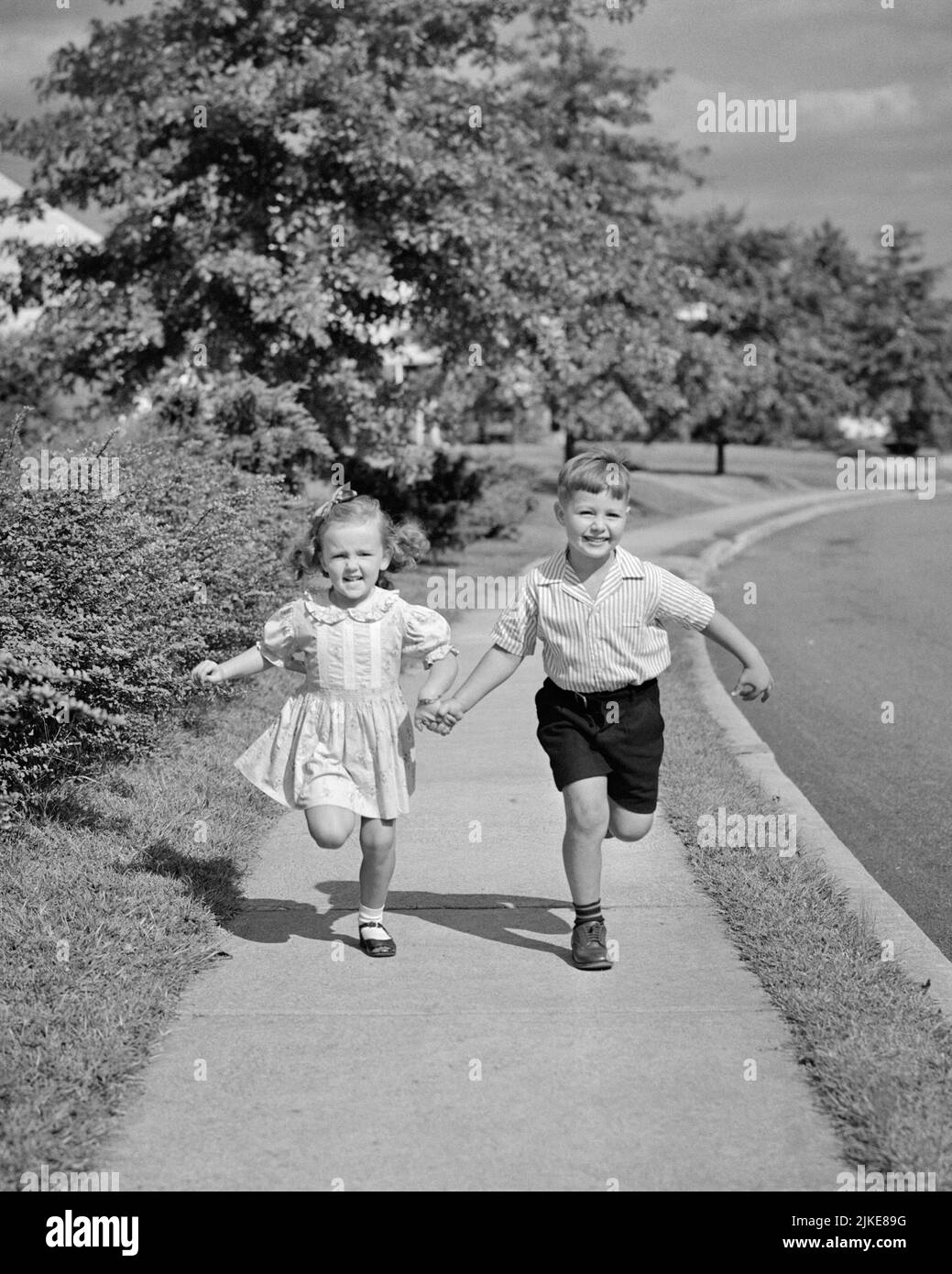 1940s 1950s BOY BROTHER AND GIRL SISTER HOLDING HANDS RUNNING DOWN SUBURBAN STREET TOWARDS LOOKING AT CAMERA - j619 HAR001 HARS FITNESS JUVENILE STYLE HEALTHY TEAMWORK ATHLETE JOY LIFESTYLE SPEED CELEBRATION FEMALES BROTHERS HEALTHINESS HOME LIFE ATHLETICS COPY SPACE FRIENDSHIP FULL-LENGTH PERSONS INSPIRATION CARING MALES RISK ATHLETIC SIBLINGS TOWARDS CONFIDENCE SISTERS B&W EYE CONTACT ACTIVITY HAPPINESS PHYSICAL WELLNESS HIGH ANGLE STRENGTH AND EXCITEMENT RECREATION SIBLING CONNECTION CONCEPTUAL ATHLETES FLEXIBILITY MUSCLES STYLISH SHORT PANTS COOPERATION GROWTH JUVENILES TOGETHERNESS Stock Photo